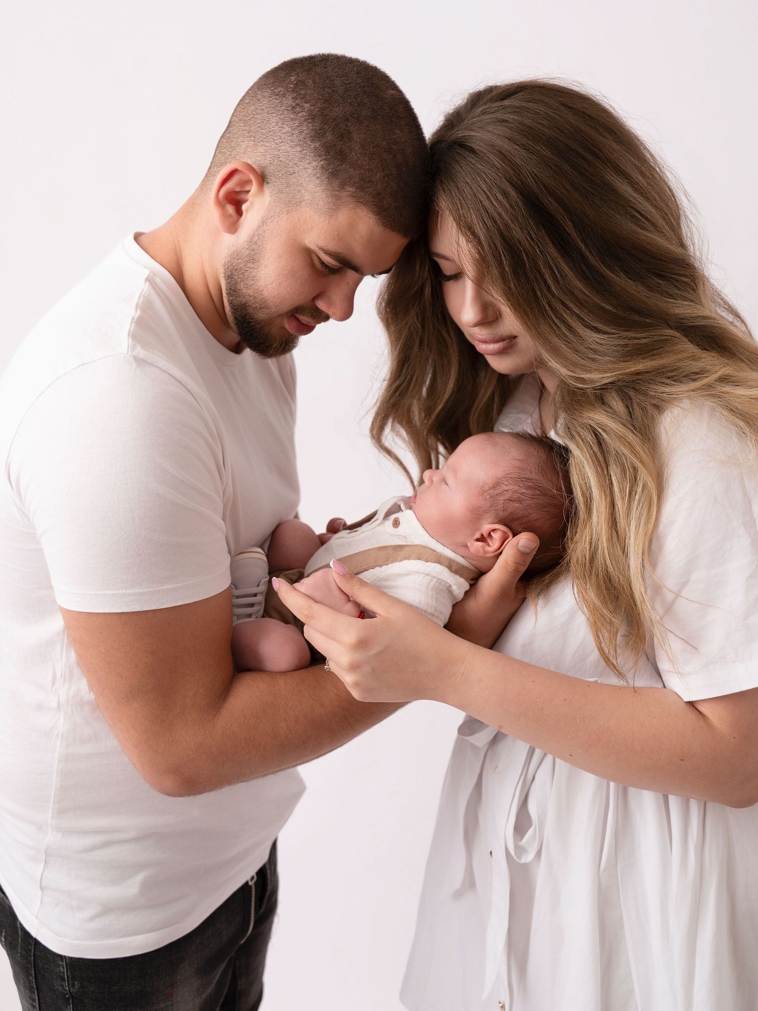 New parents in white outfits tenderly hold their sleeping newborn baby together in a bright studio portrait.