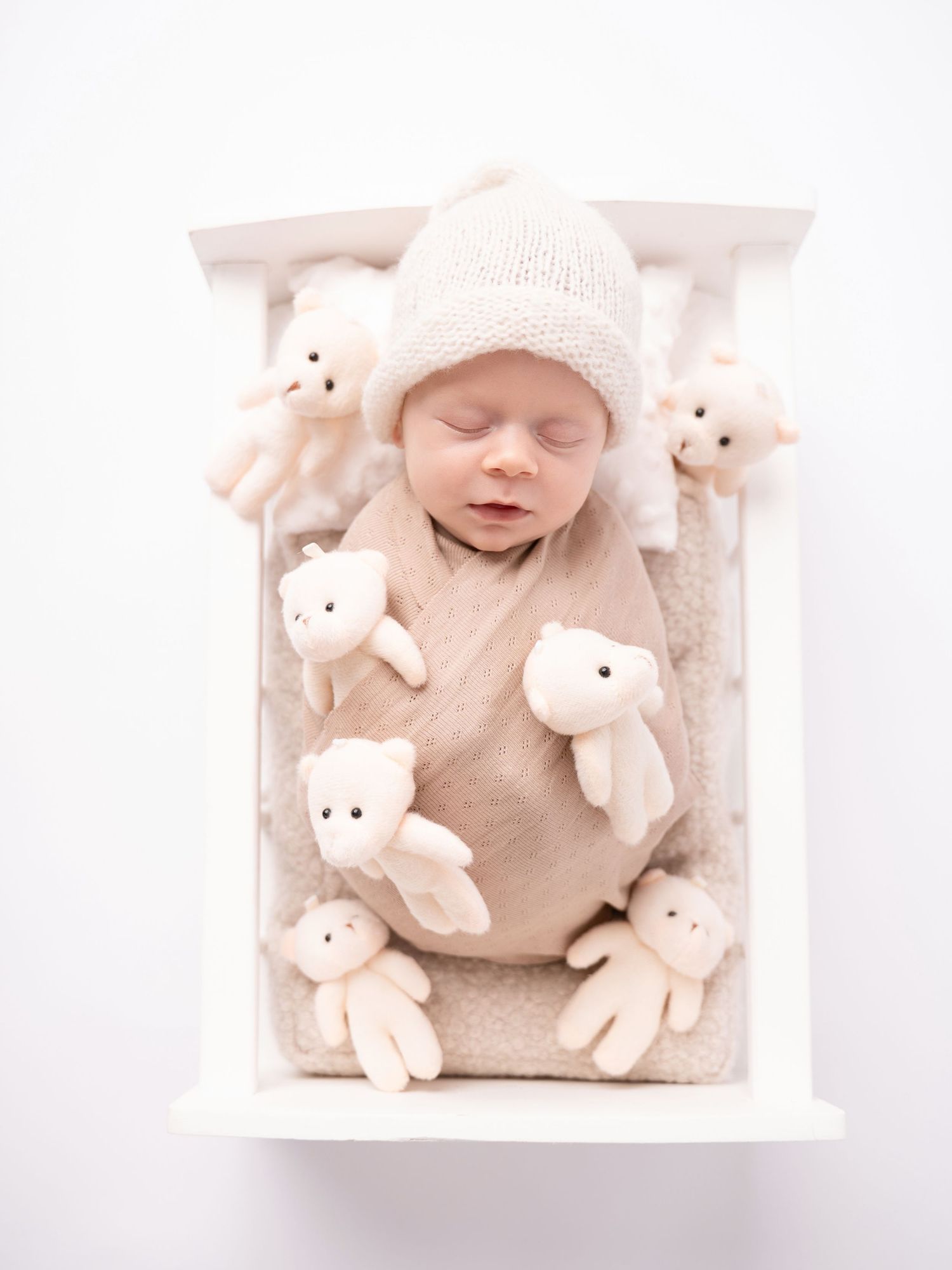 Sleeping newborn baby in a beige knit hat and swaddle surrounded by white teddy bears in a small white crib.