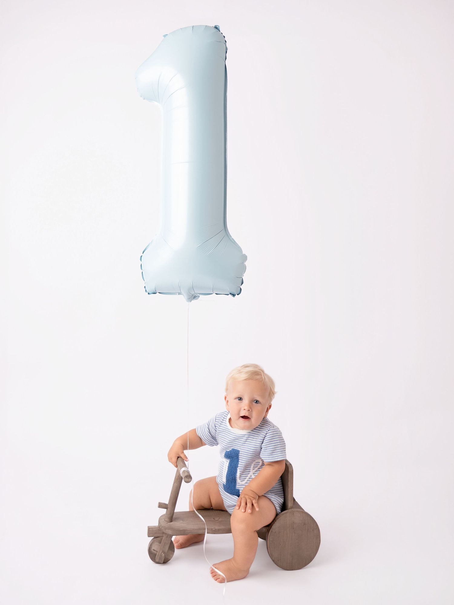 Baby boy sitting on wooden toy bike with blue number 1 balloon for first birthday photo session.