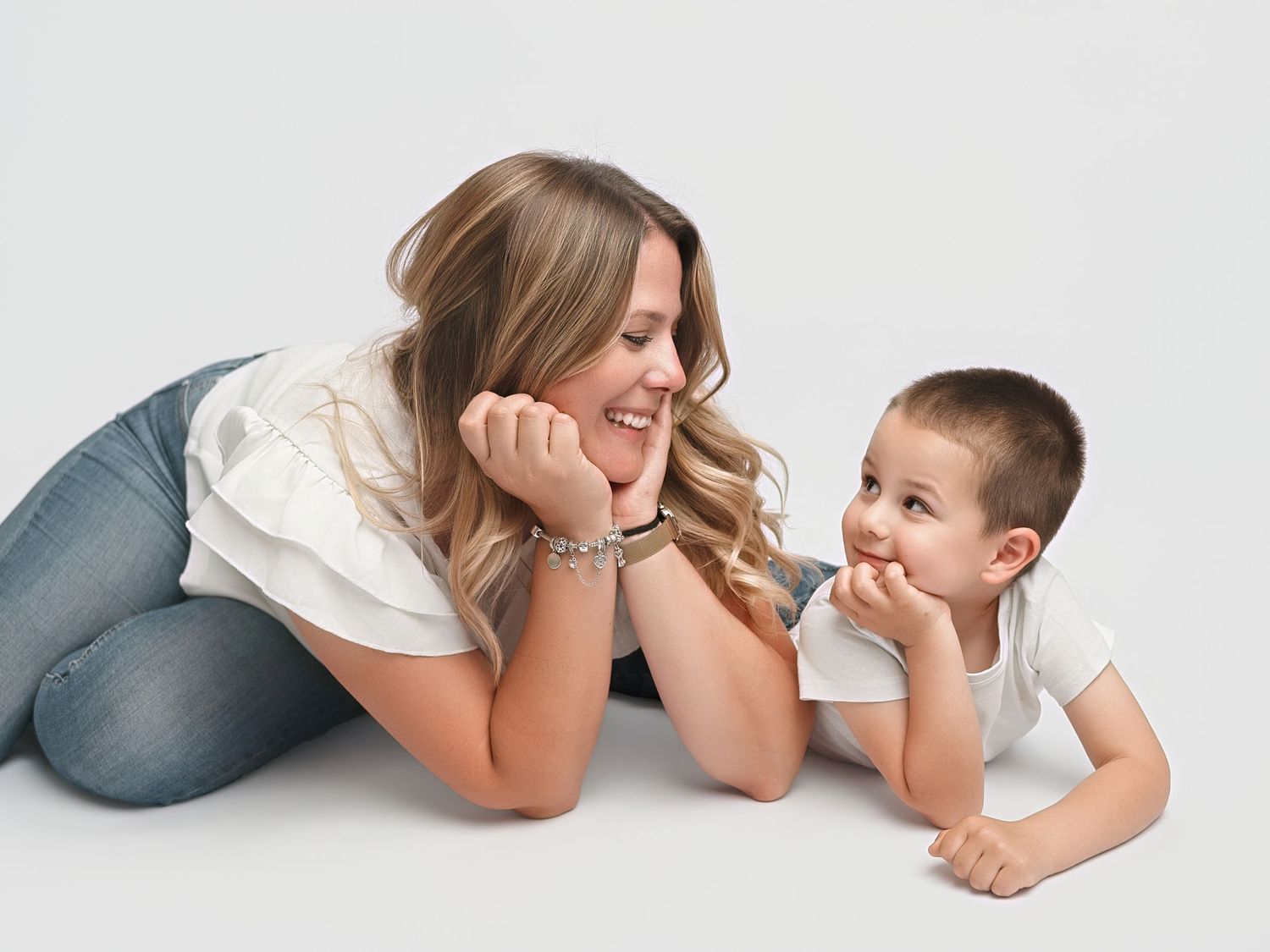 A smiling mother and her young child lie on the floor face-to-face, sharing a playful and loving moment together.
