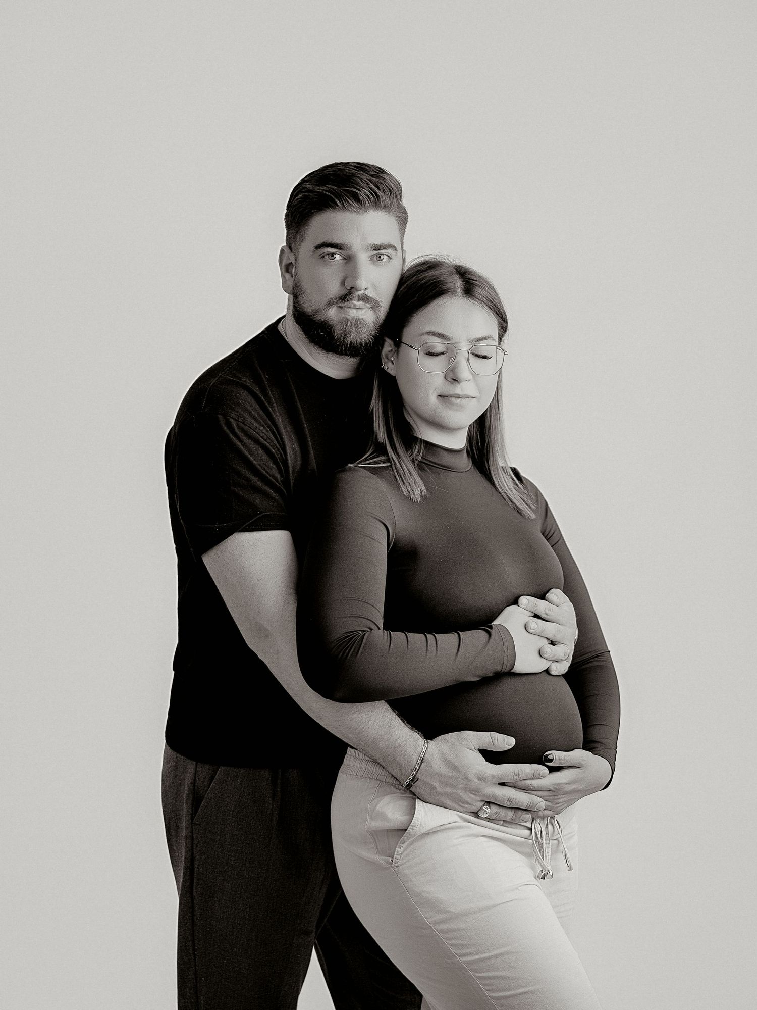 Couple embracing in black and white maternity portrait, man holding pregnant woman from behind on light background.