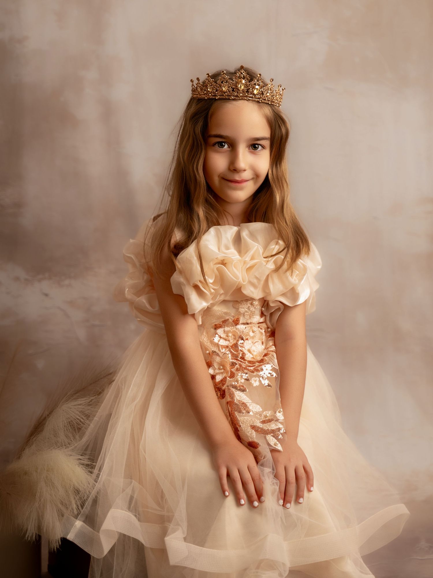 Young girl in elegant cream tulle dress and gold crown poses gracefully for a princess-themed portrait photo.