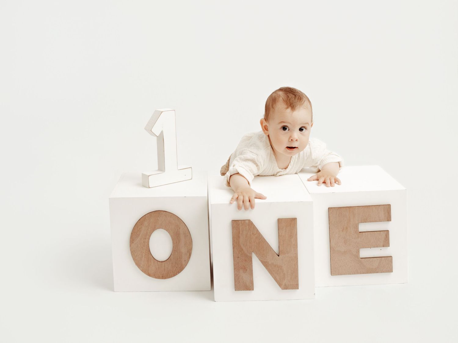 Curious baby peeking over white wooden blocks spelling ONE for first birthday photos.