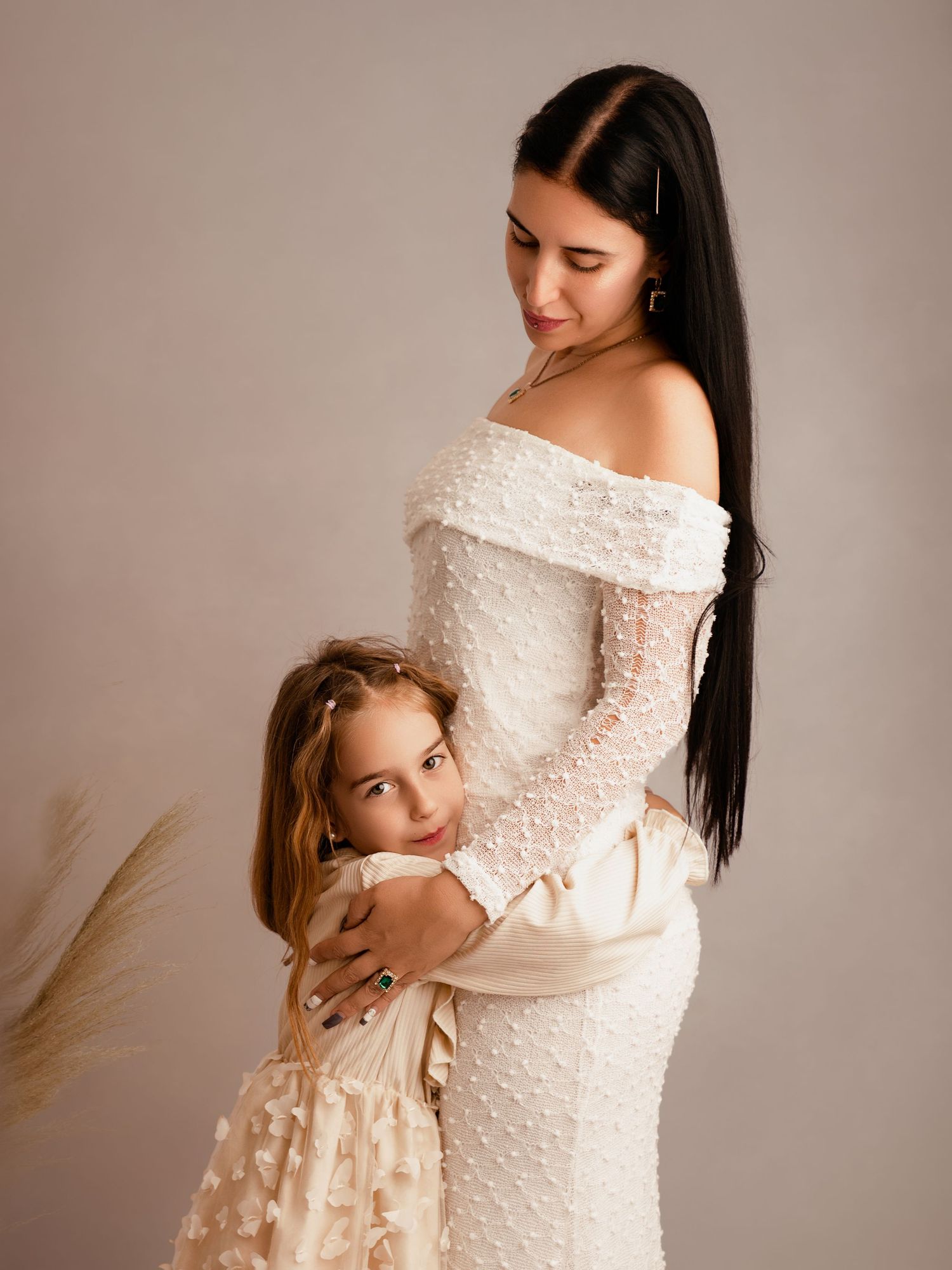 Pregnant woman in white pearl dress embracing young daughter in cream floral gown during maternity photo shoot.