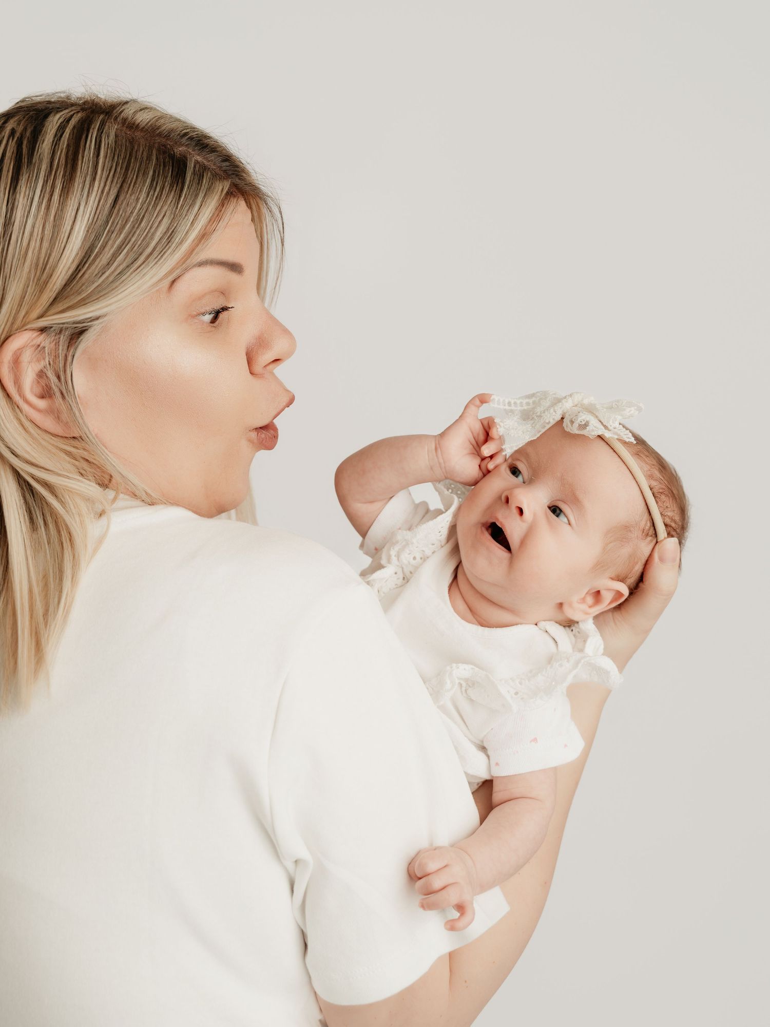 Blonde mother making funny face at wide-eyed newborn baby in white outfit against light background.