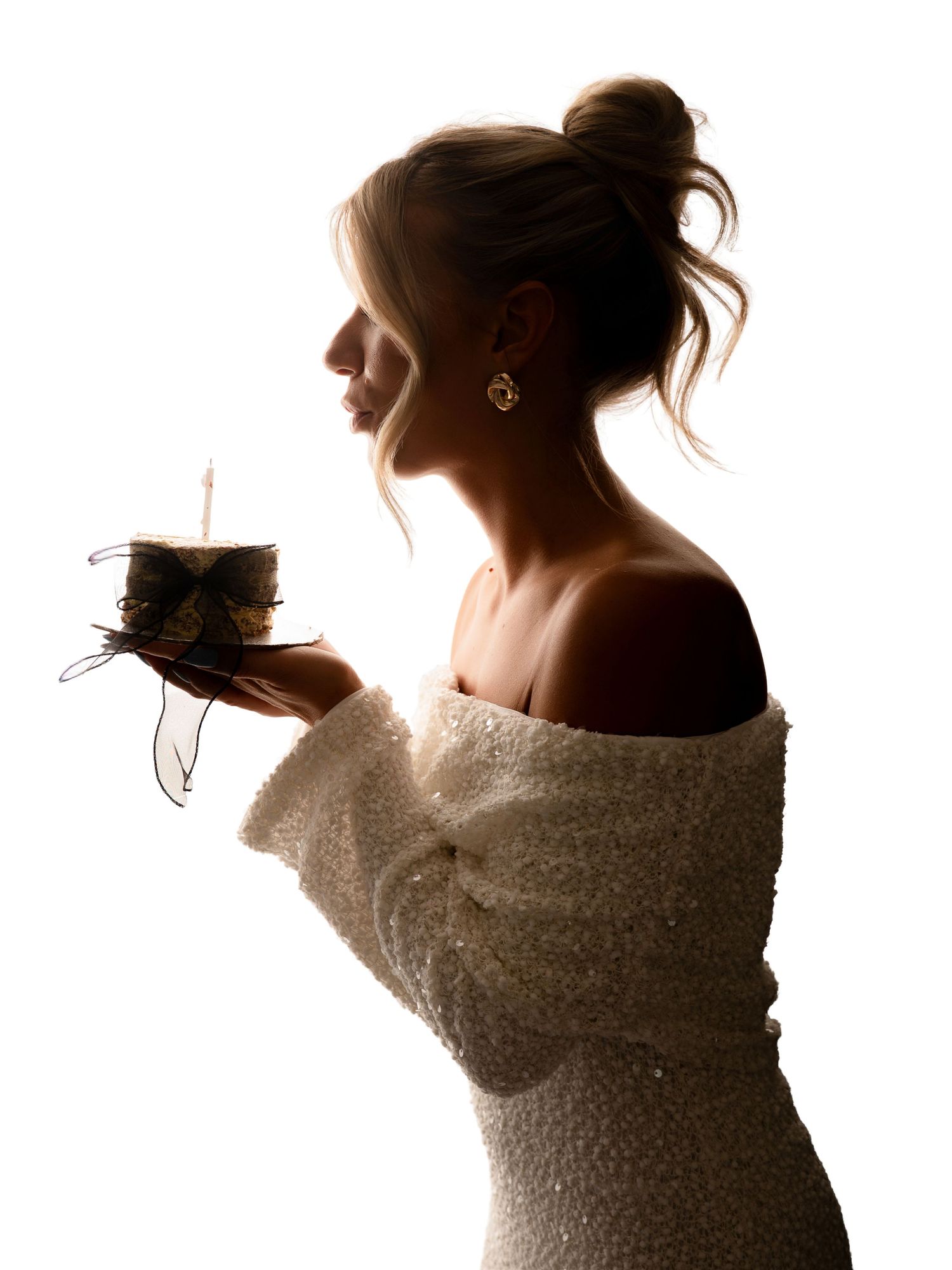 Silhouette profile of woman in white off-shoulder dress holding a small birthday cake with candle.