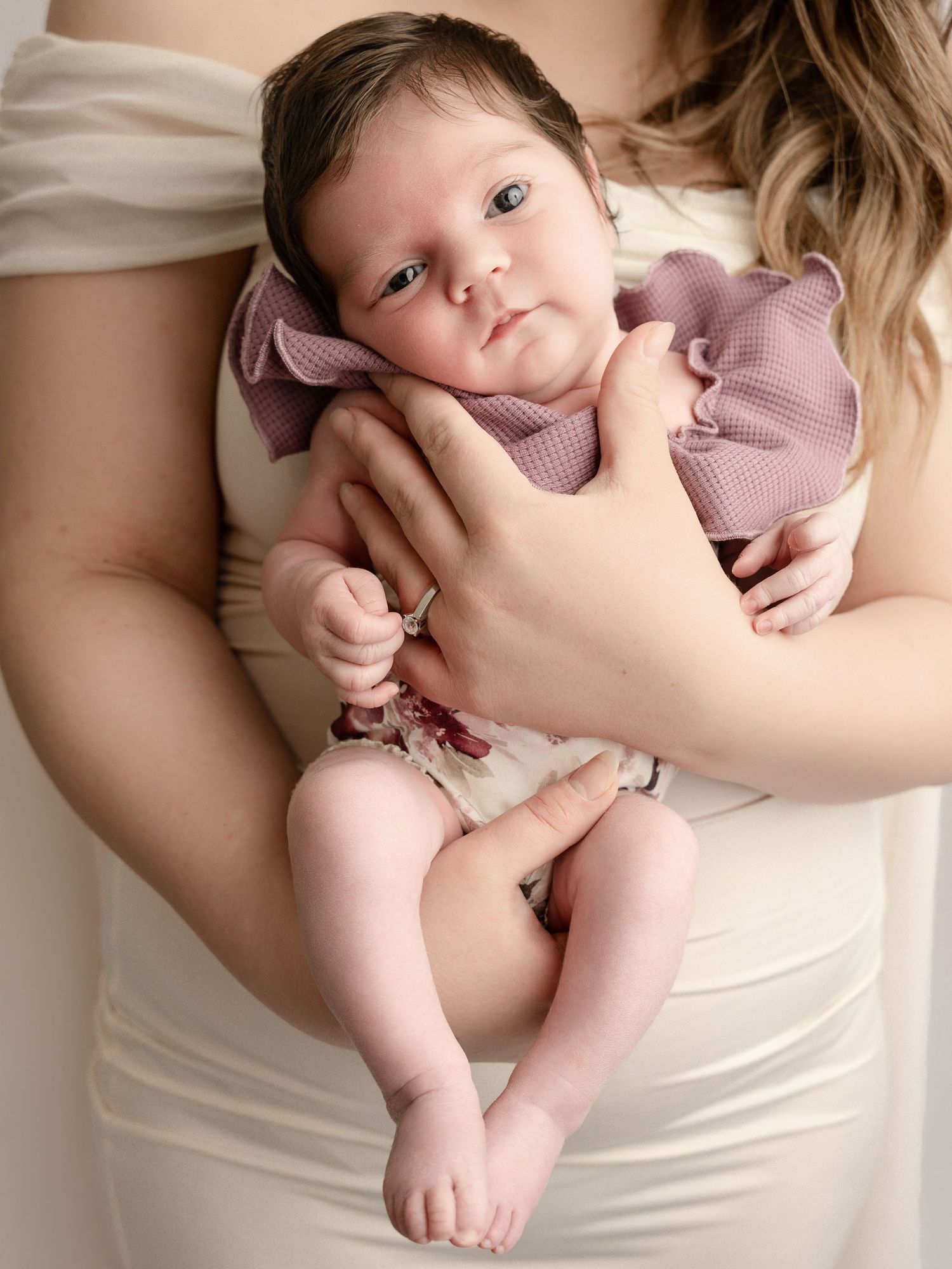 Close-up of newborn baby in mauve ruffled romper being cradled in mothers hands against cream dress.