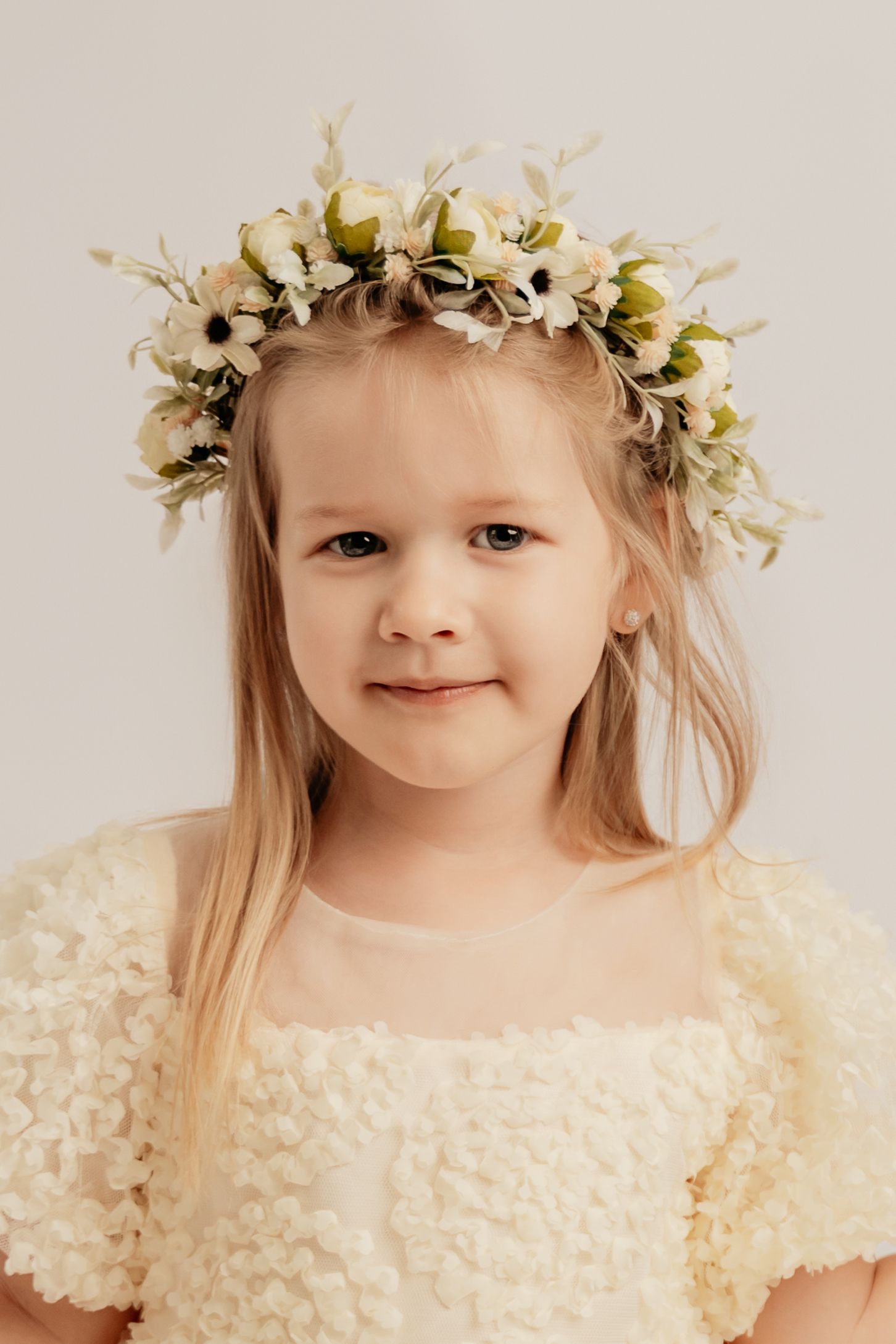 A sweet young blonde girl wearing a white floral crown and cream textured dress smiles softly at camera.