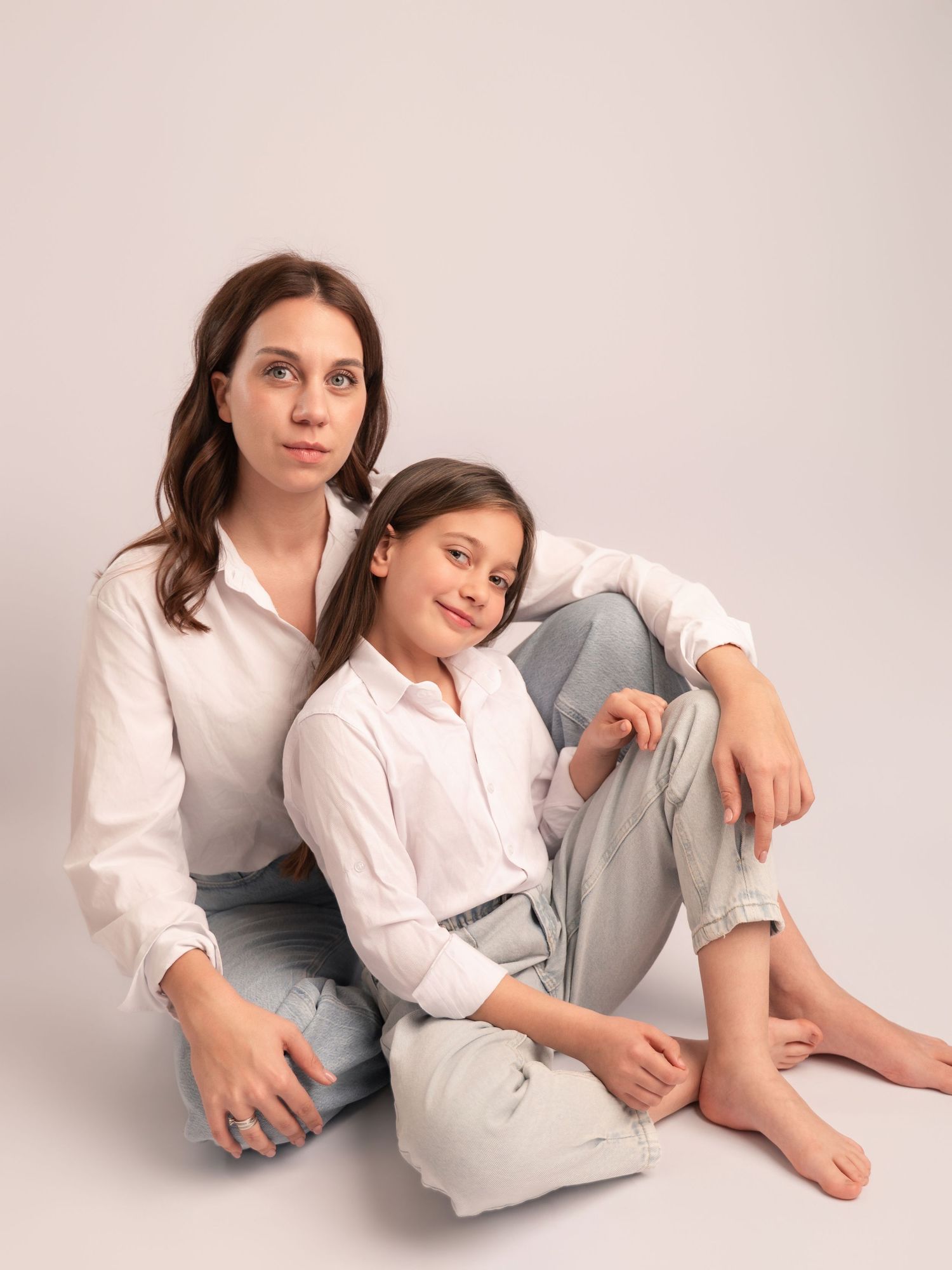 A mother and daughter in white shirts and jeans sit together in a relaxed studio portrait pose.