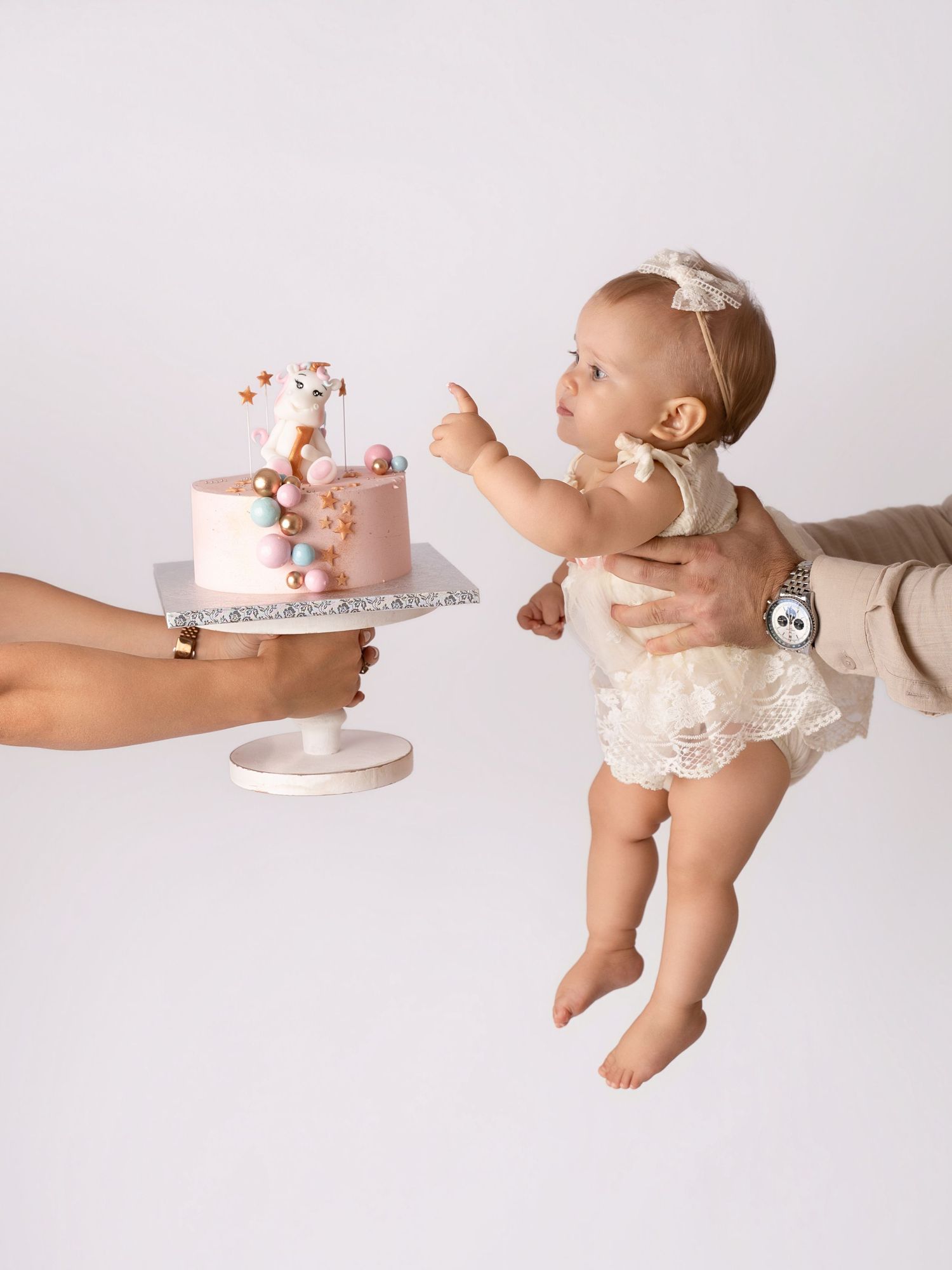 Baby girl in lace outfit reaches for pink giraffe birthday cake held by adult hands in studio photo.