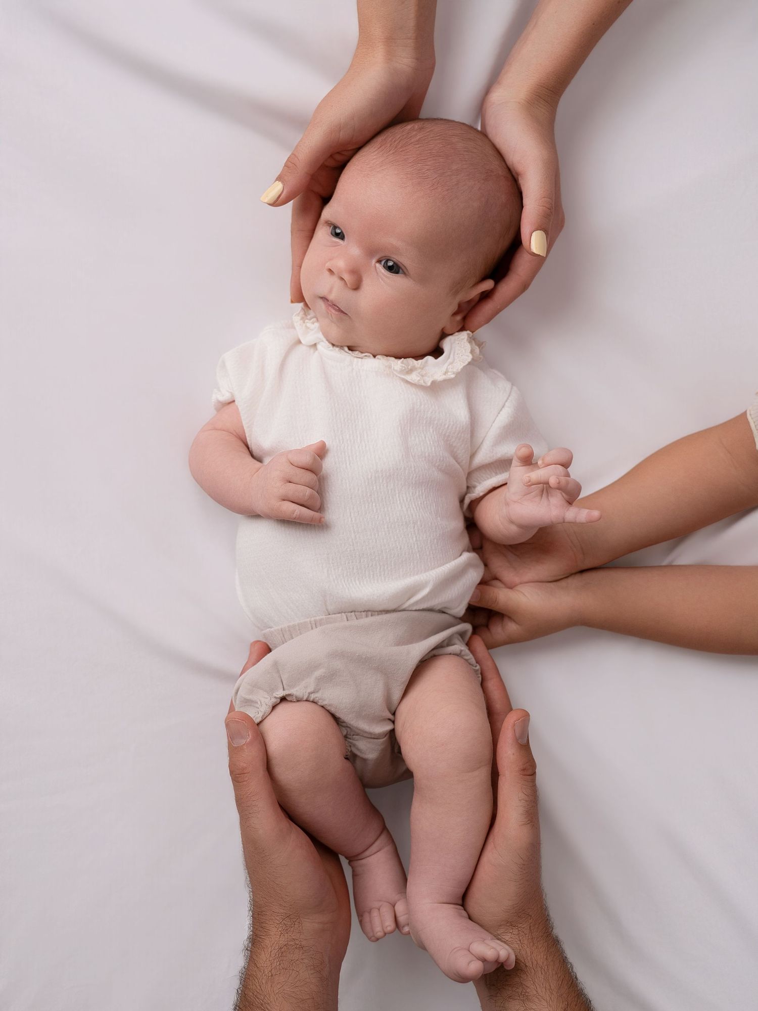 Newborn baby in white onesie cradled by two pairs of adult hands on a soft white background.