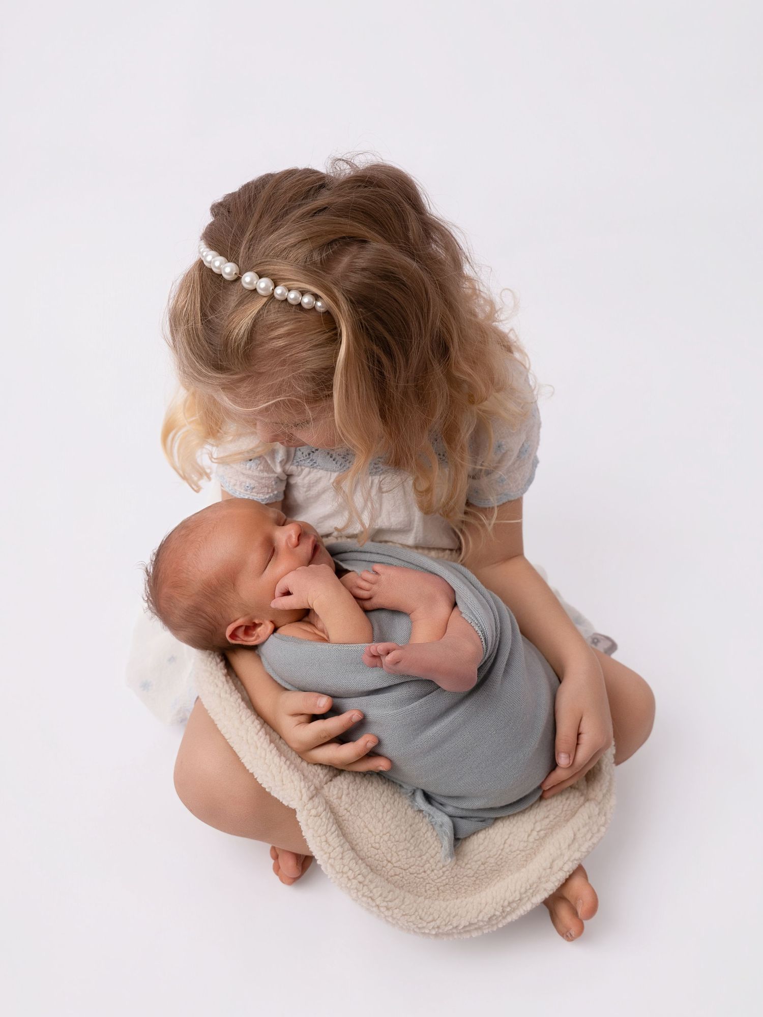 Young girl with pearl headband gently holds sleeping newborn baby wrapped in blue on white background.