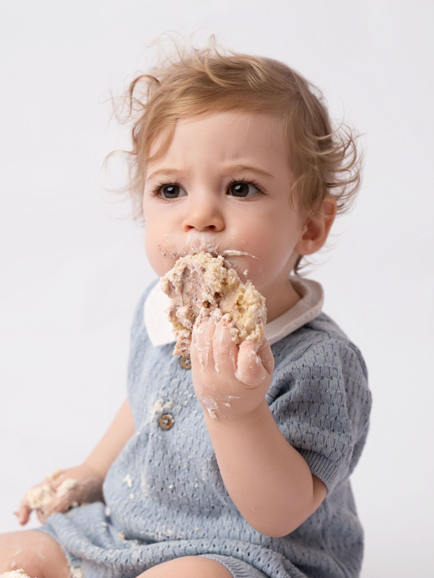 Baby in blue knit outfit eating birthday smash cake with messy hands and face.