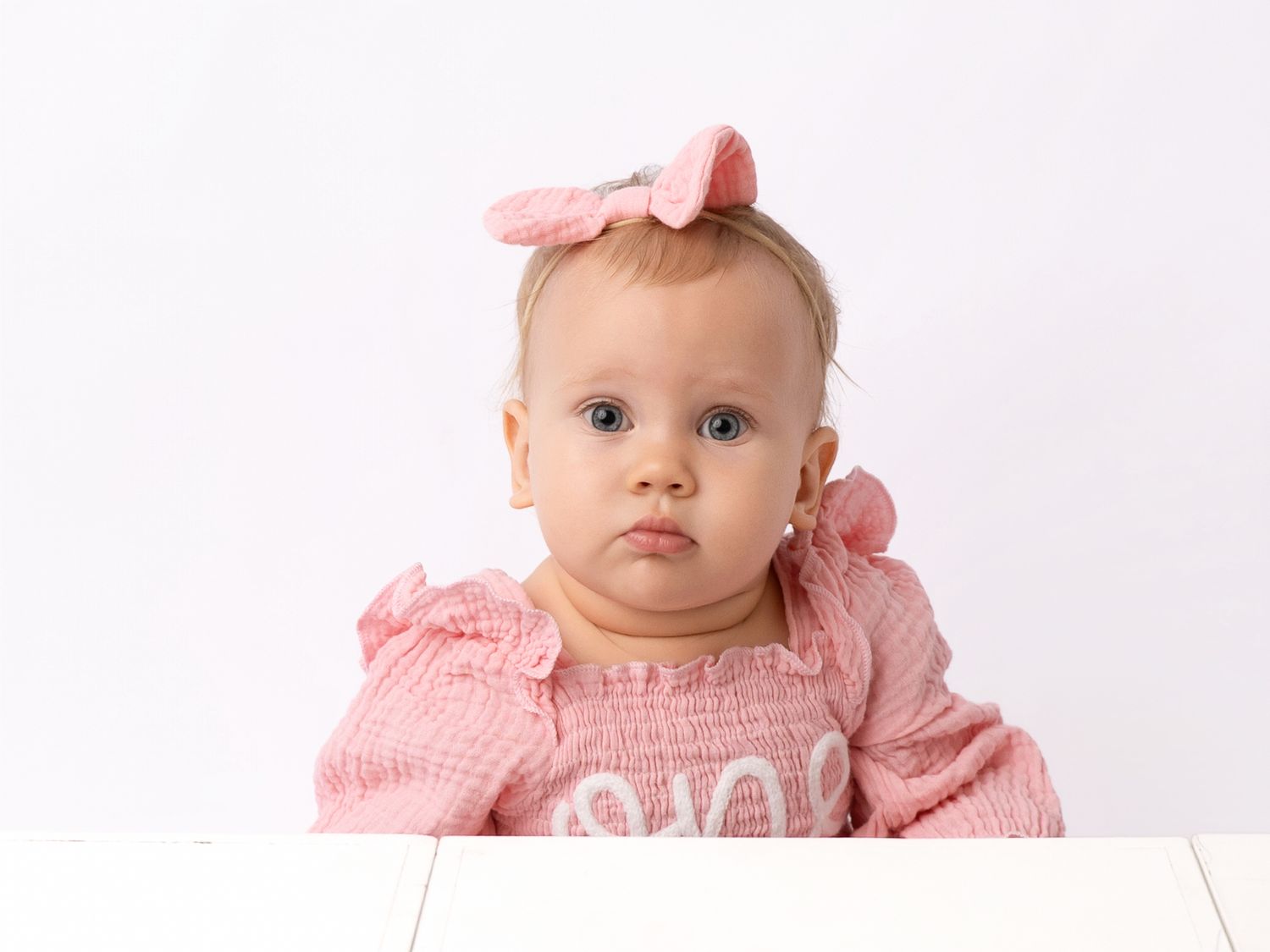 Adorable baby girl in pink outfit and bow headband poses for a professional white background portrait photo.