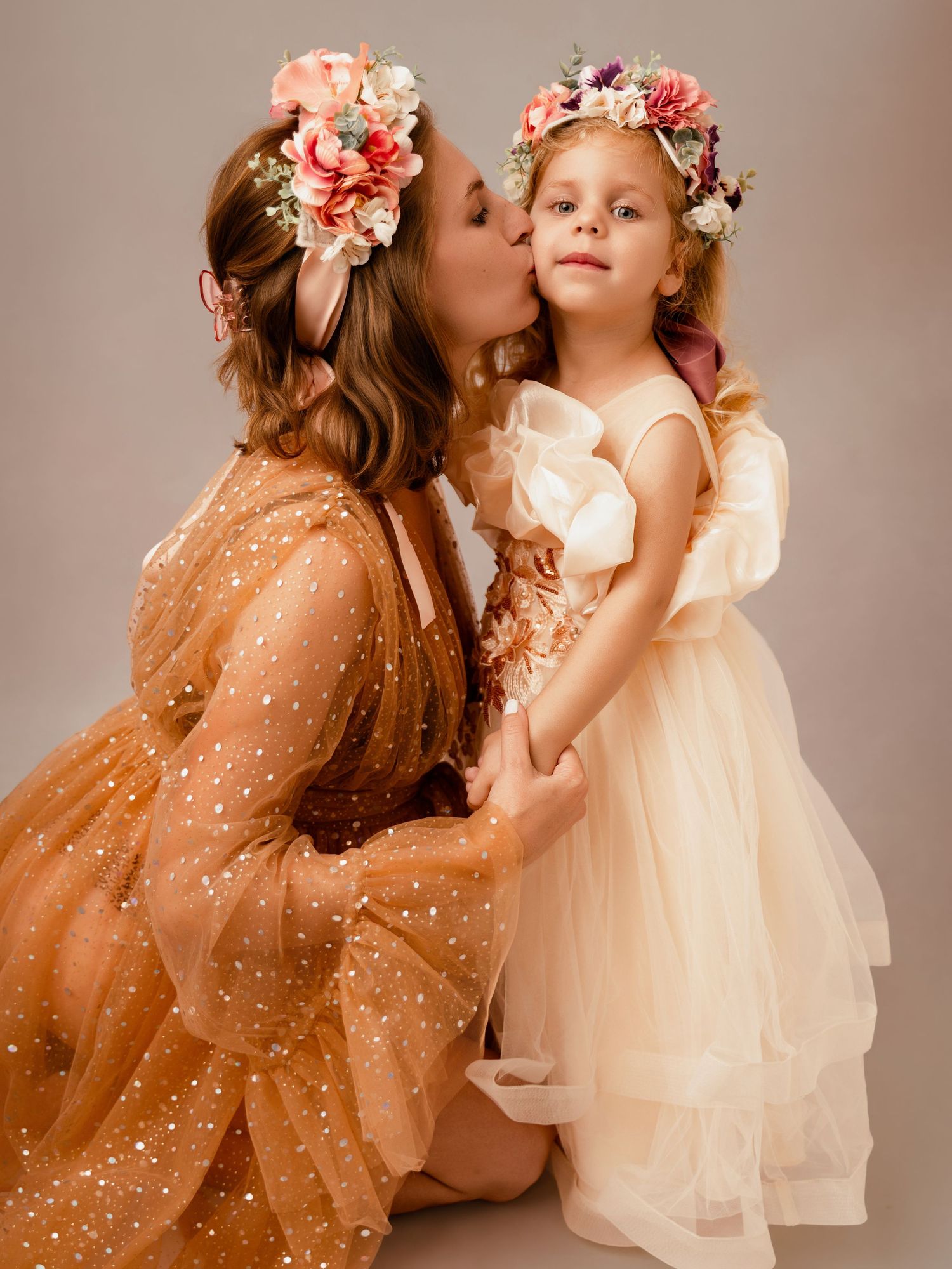 Woman in gold tulle dress kisses girl in cream gown, both wearing floral crowns, in a studio portrait.