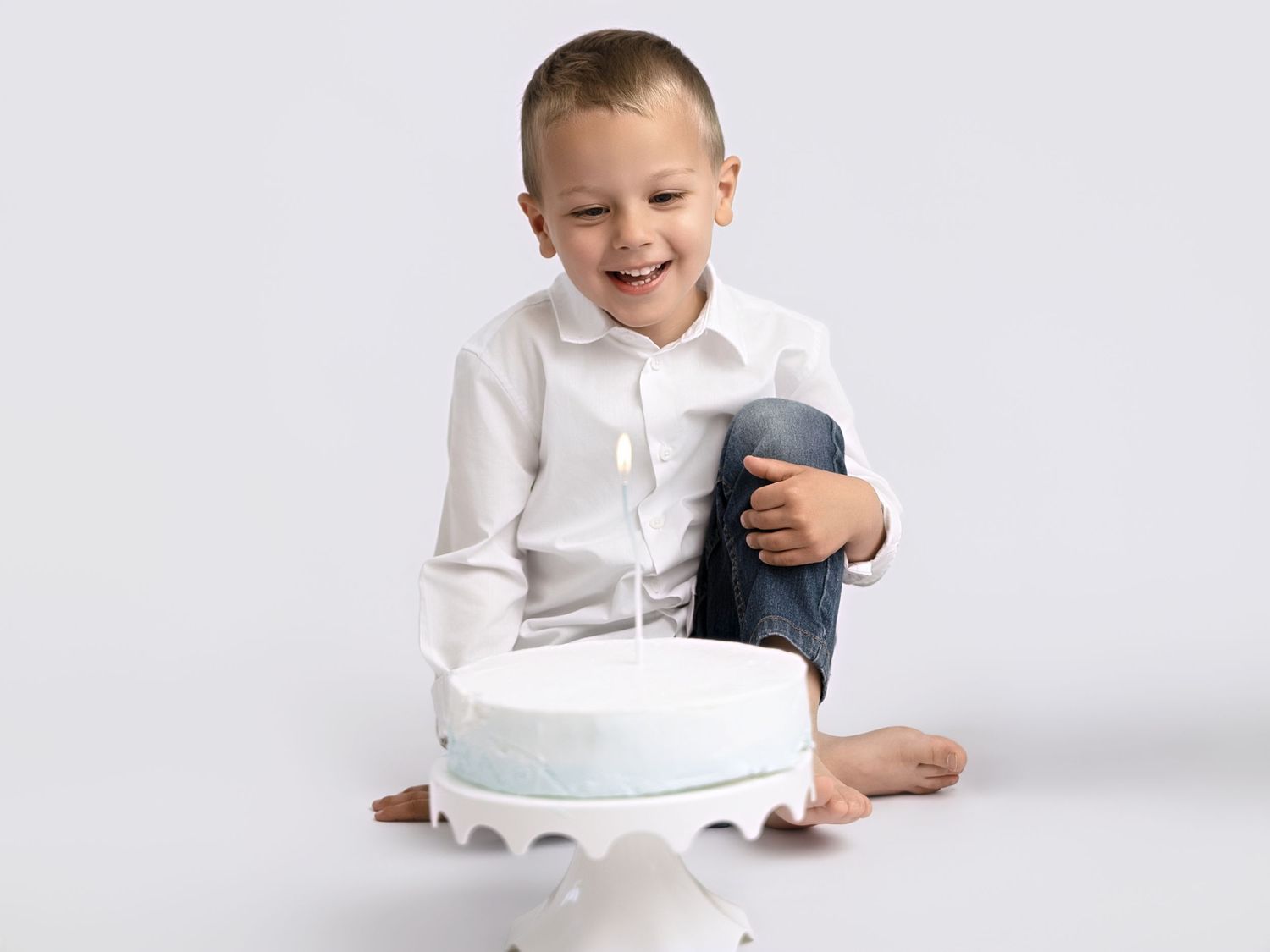 Happy young boy in white shirt and jeans sitting cross-legged with a white birthday cake with one candle.