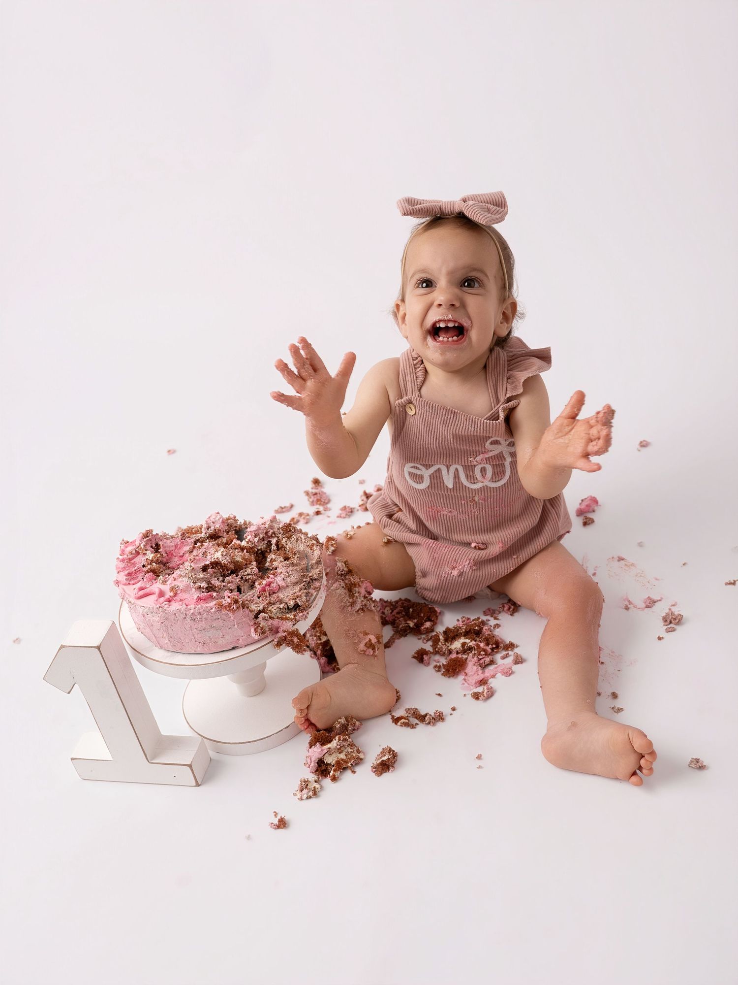 Baby girl in pink romper joyfully smashing her first birthday cake during a cake smash photo session.