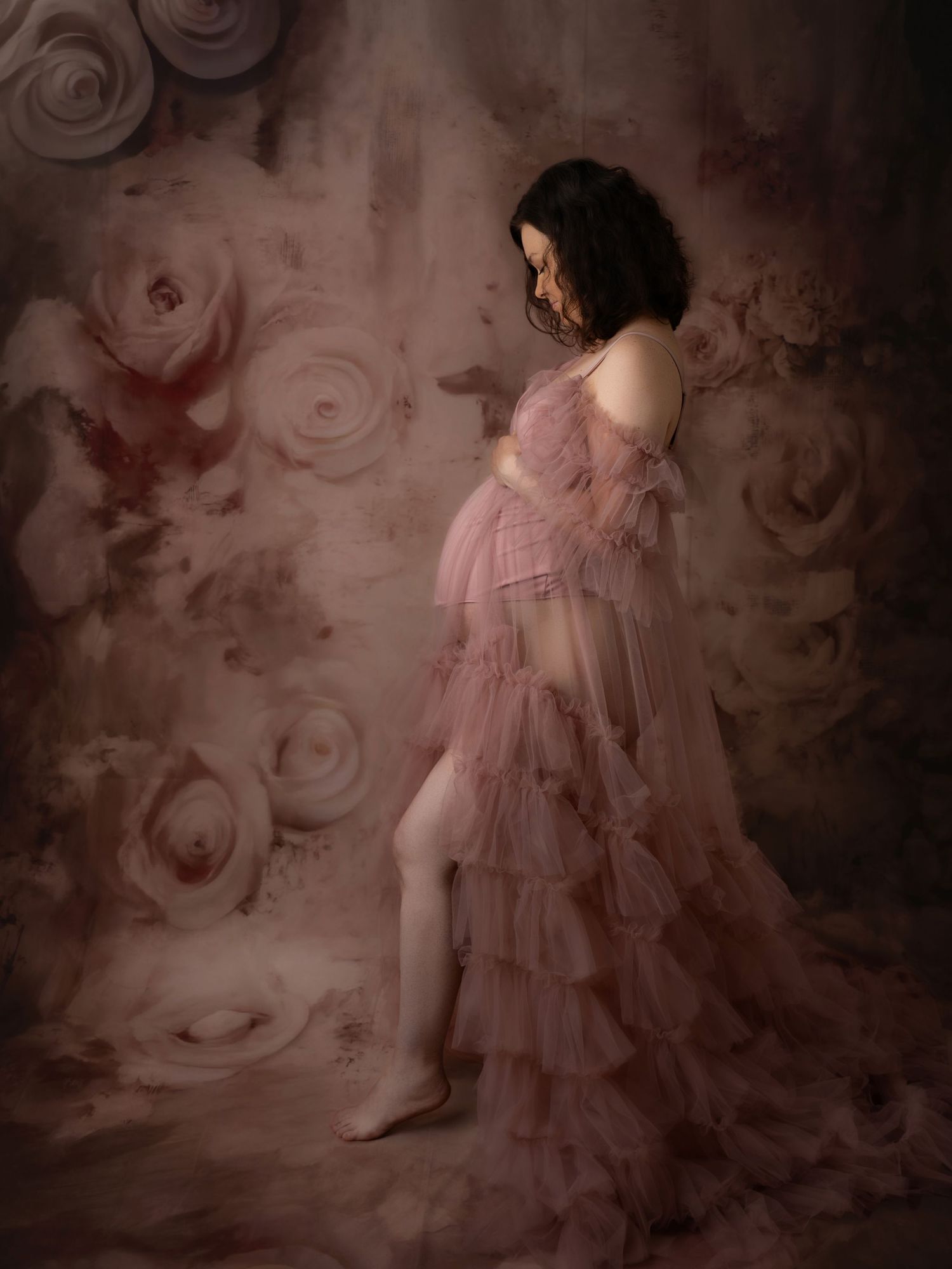Pregnant woman in pink tulle maternity gown posing against floral painted backdrop in professional photo session.