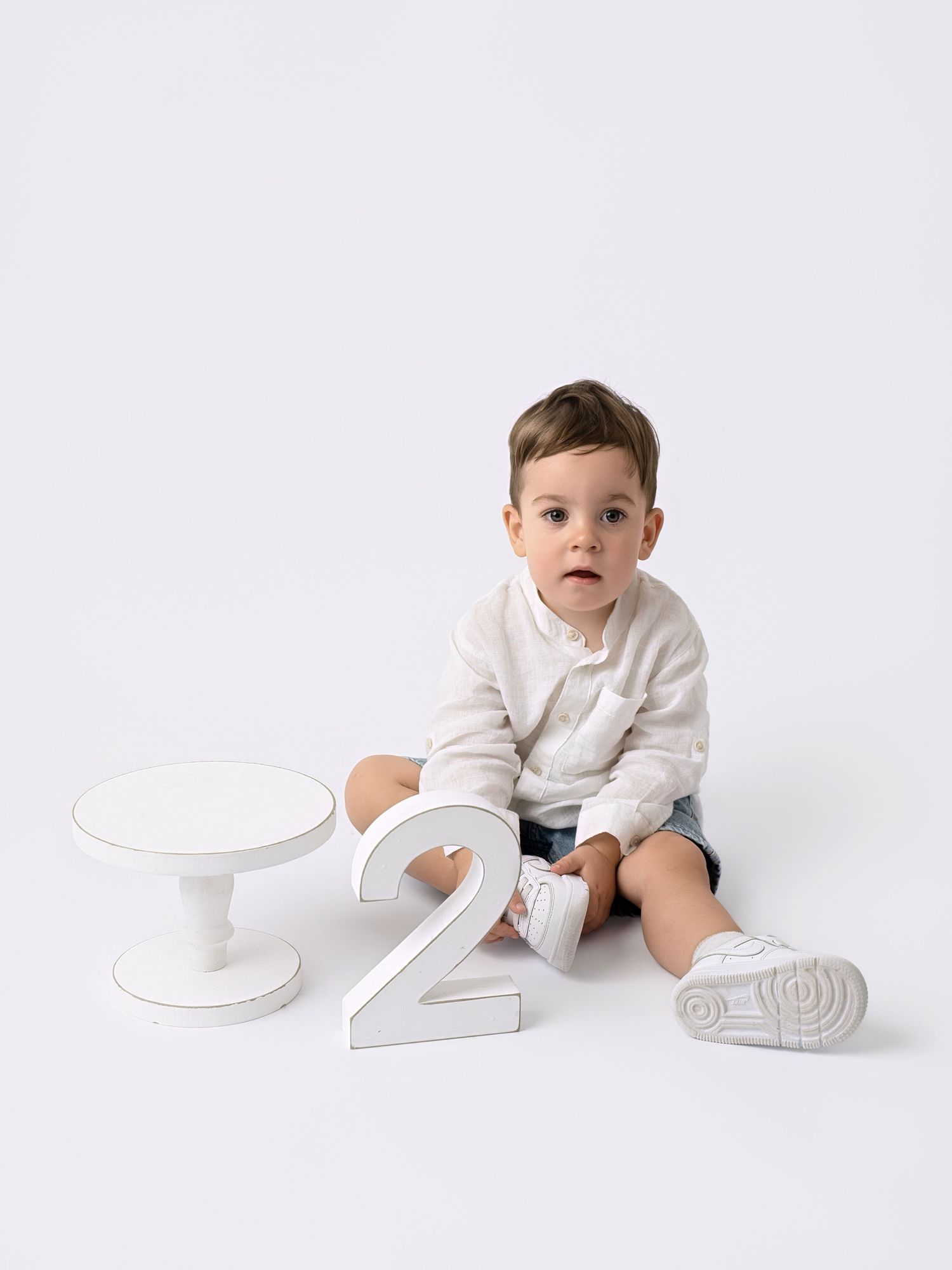 Toddler boy in white shirt and denim shorts sits beside a number 2 prop for a 2nd birthday photo session.