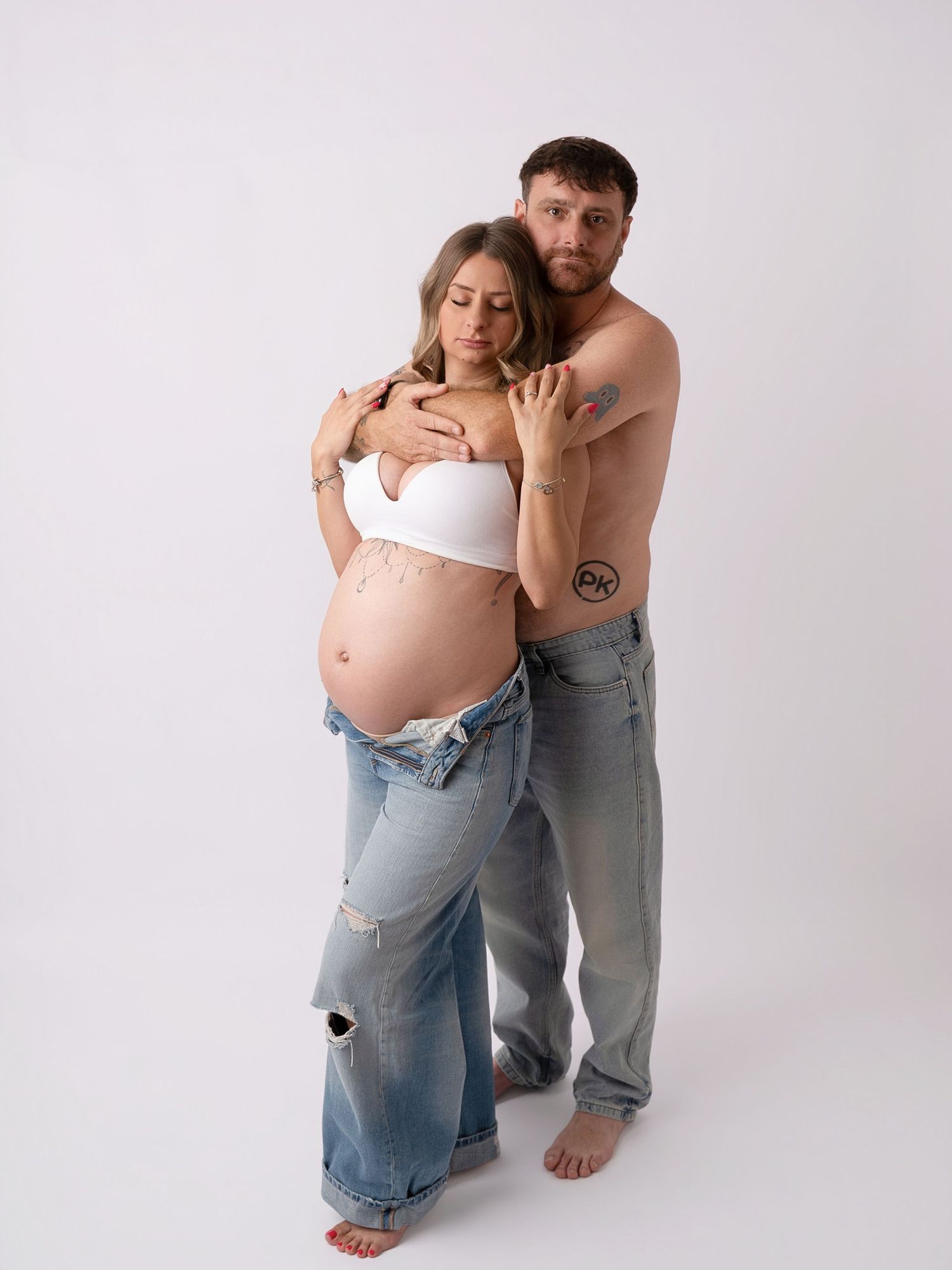 Couple maternity photo with shirtless man embracing pregnant woman in white bralette and low-rise jeans on white background.