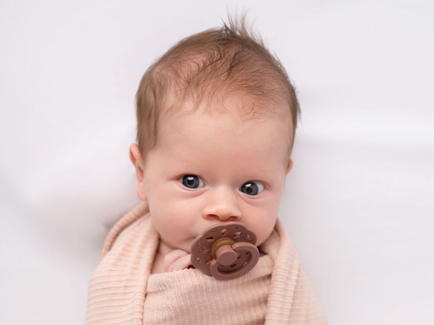 Newborn baby with blue eyes sucking a brown pacifier, wrapped in a soft pink knit blanket on white background.
