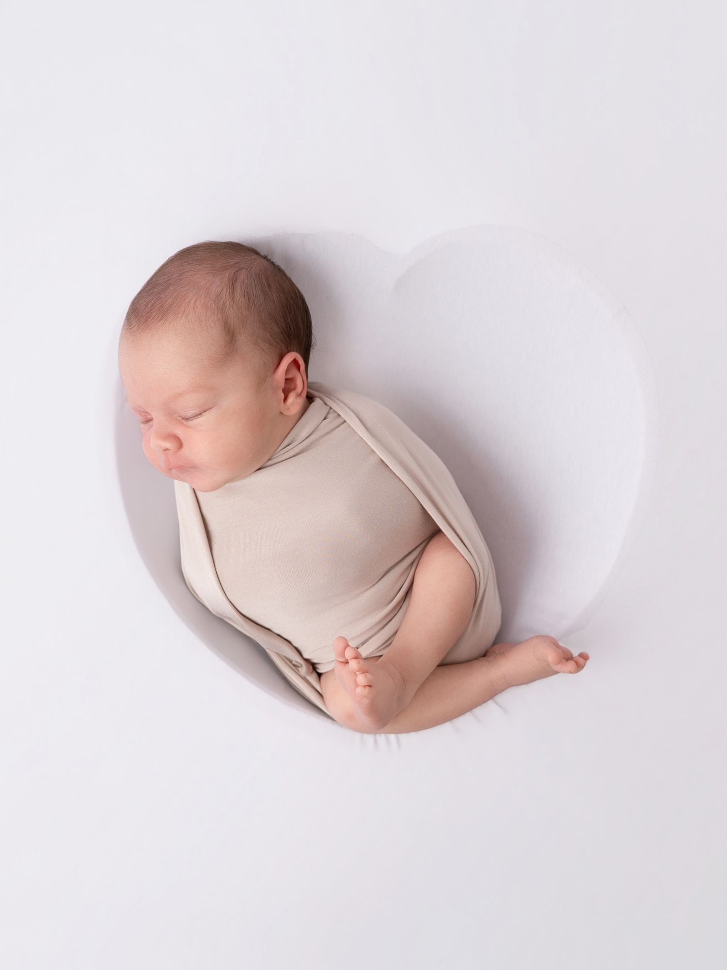 Sleeping newborn baby swaddled in beige wrap, curled inside a heart-shaped white prop for newborn photography.