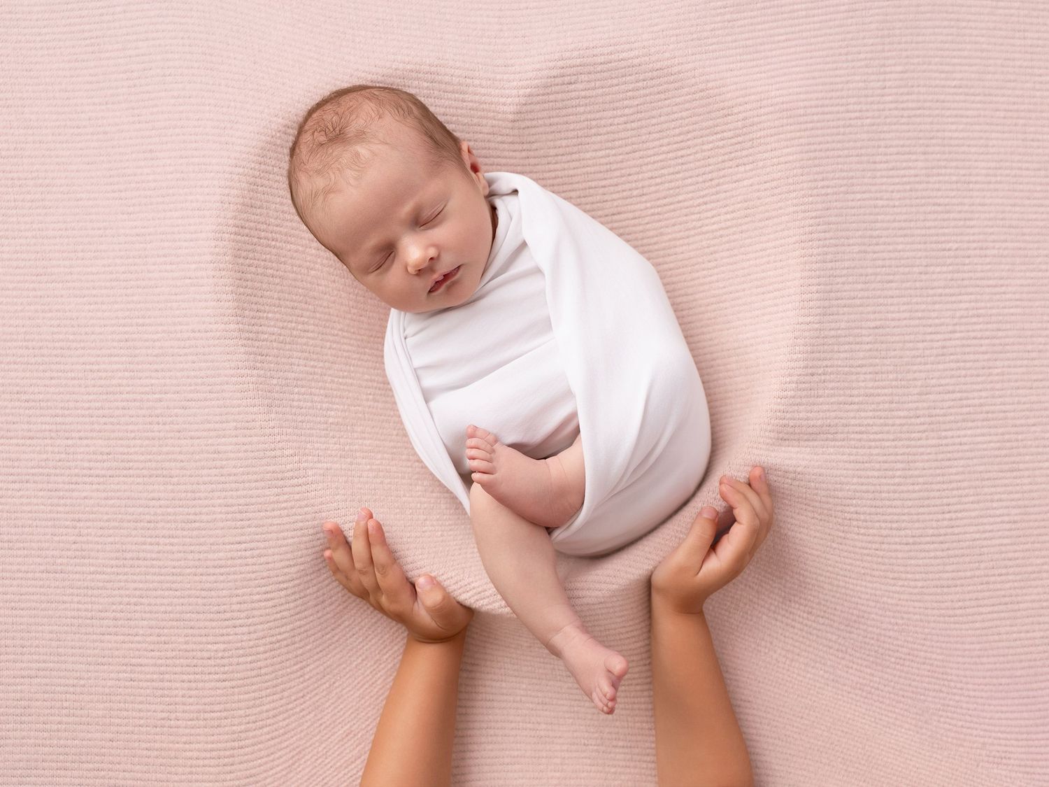 Newborn baby swaddled in white wrap resting peacefully on pink blanket, cradled by loving hands.