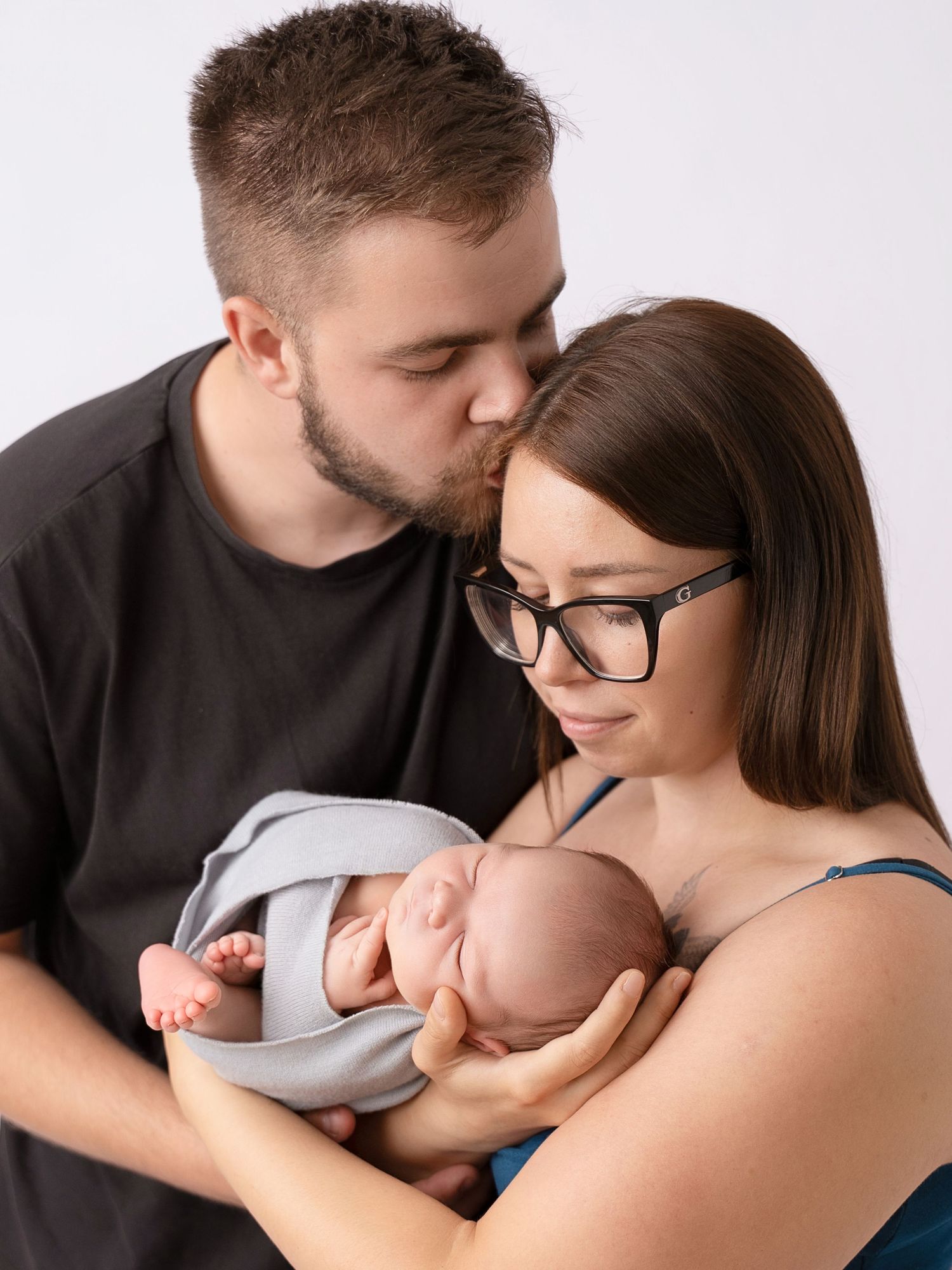 New parents lovingly holding their swaddled newborn baby, father kissing mothers head in professional studio portrait.