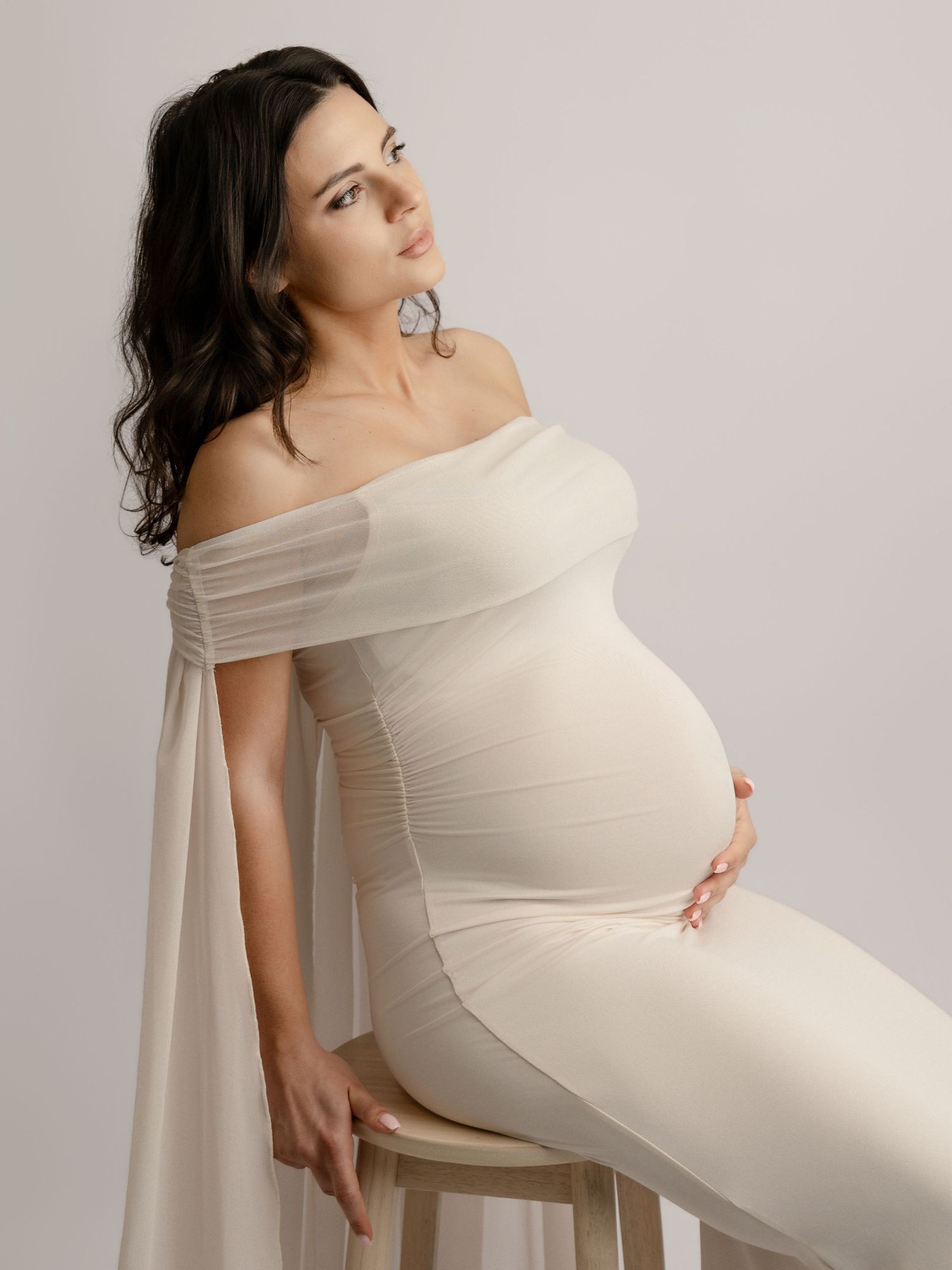 Pregnant woman in off-shoulder white fitted gown seated on stool.