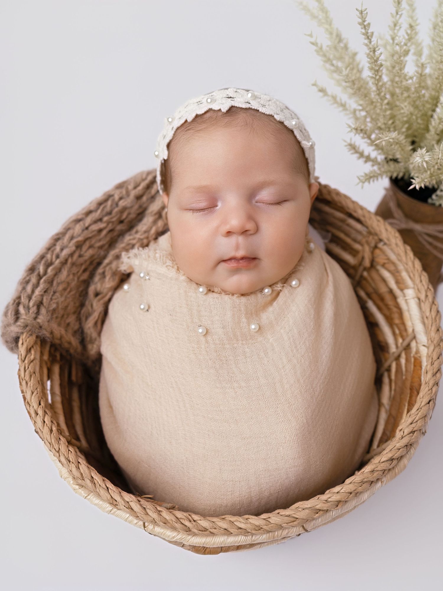 Close-up of sleeping newborn swaddled in beige wrap with pearls, wearing white lace bonnet, nestled in a wicker basket.