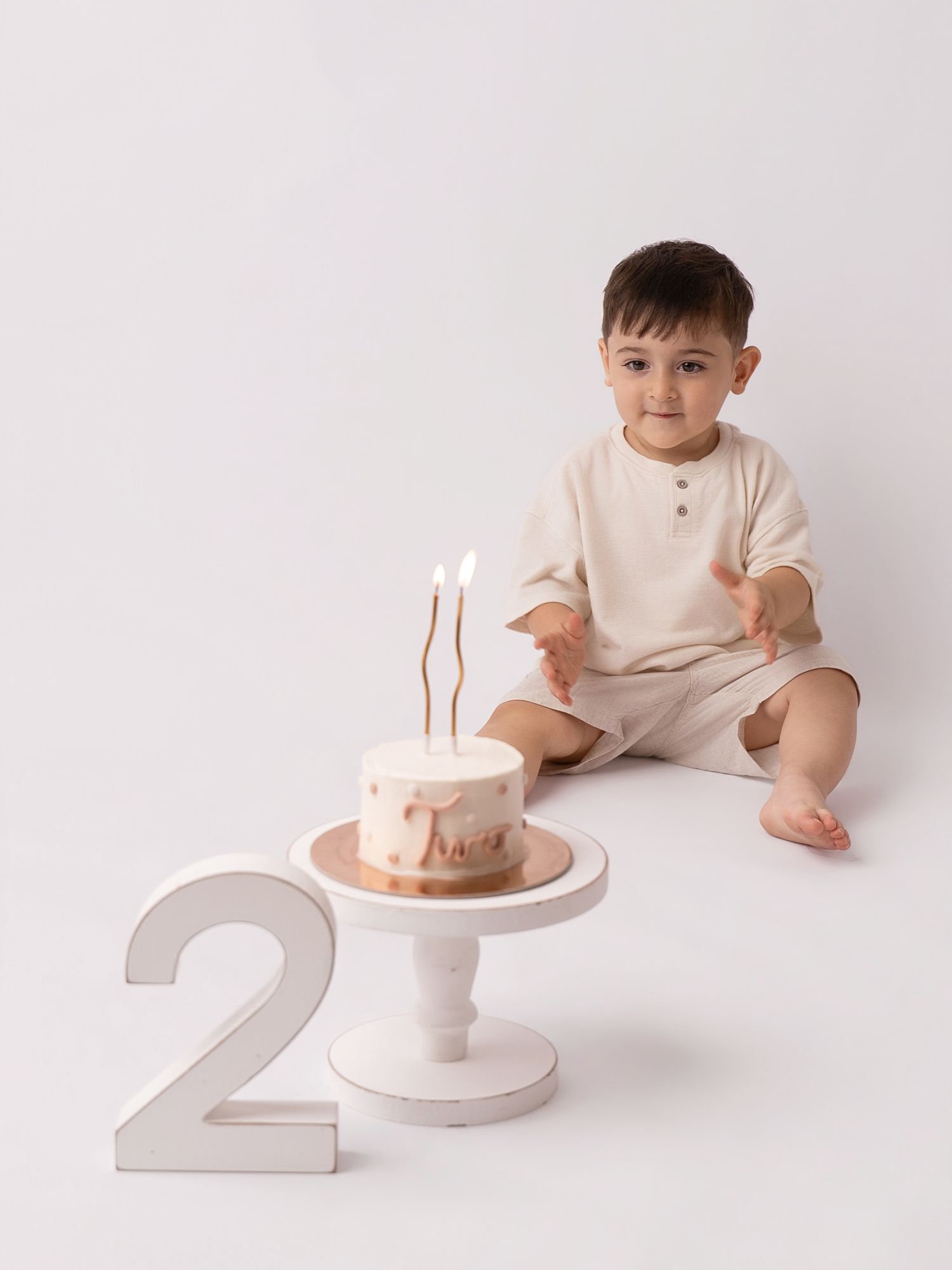 Toddler boy in cream outfit sits beside a birthday cake on white stand with number 2, celebrating his second birthday.