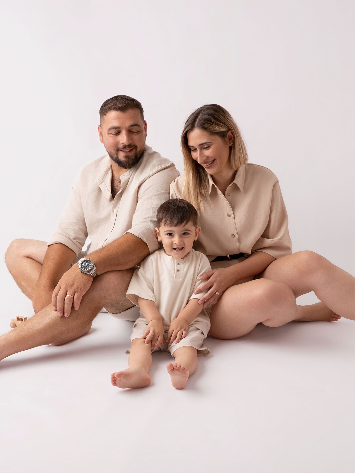 Family of three in matching beige outfits sitting on white studio floor, parents smiling at toddler boy.