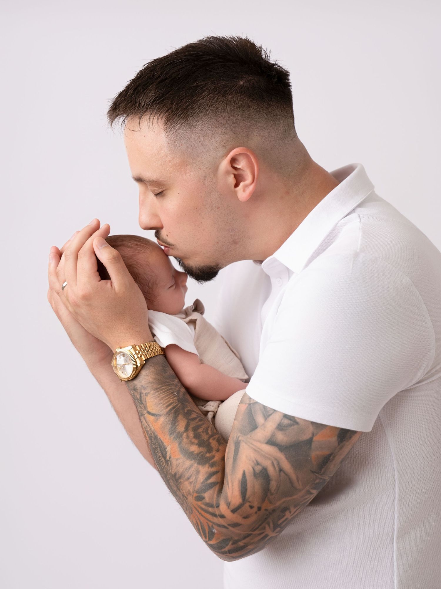 Tattooed father in white polo shirt gently kissing his newborn baby while cradling it in his arms.