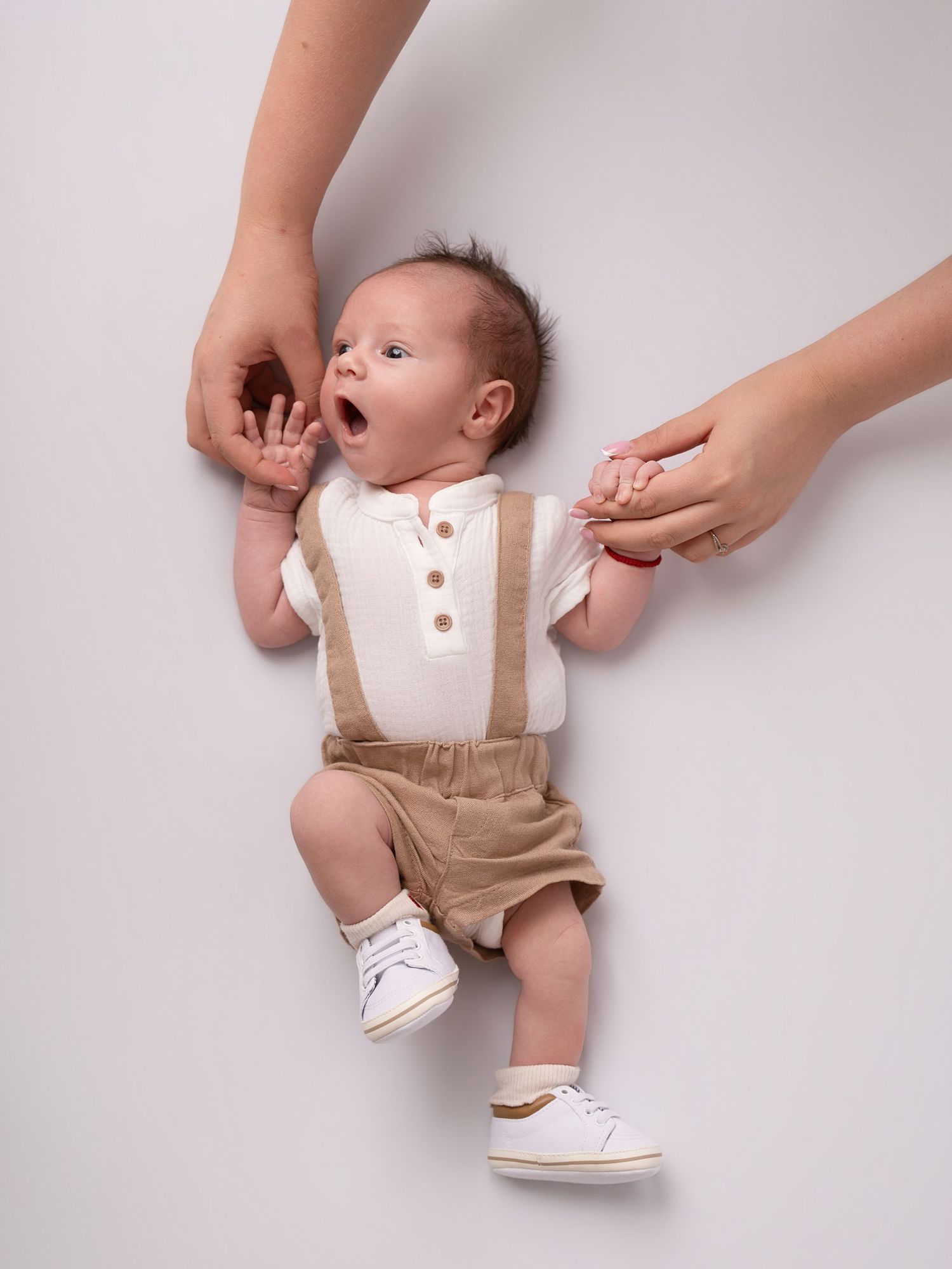 Adorable newborn baby yawning in white shirt and tan suspender shorts, held by parents hands on white background.