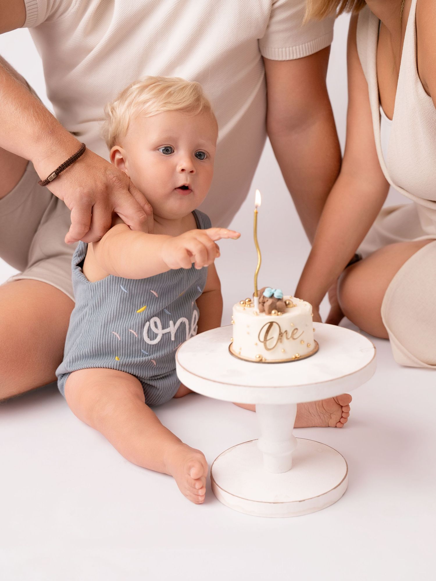 Baby's first birthday smash cake session with parents and a white cake reading One on a pedestal.