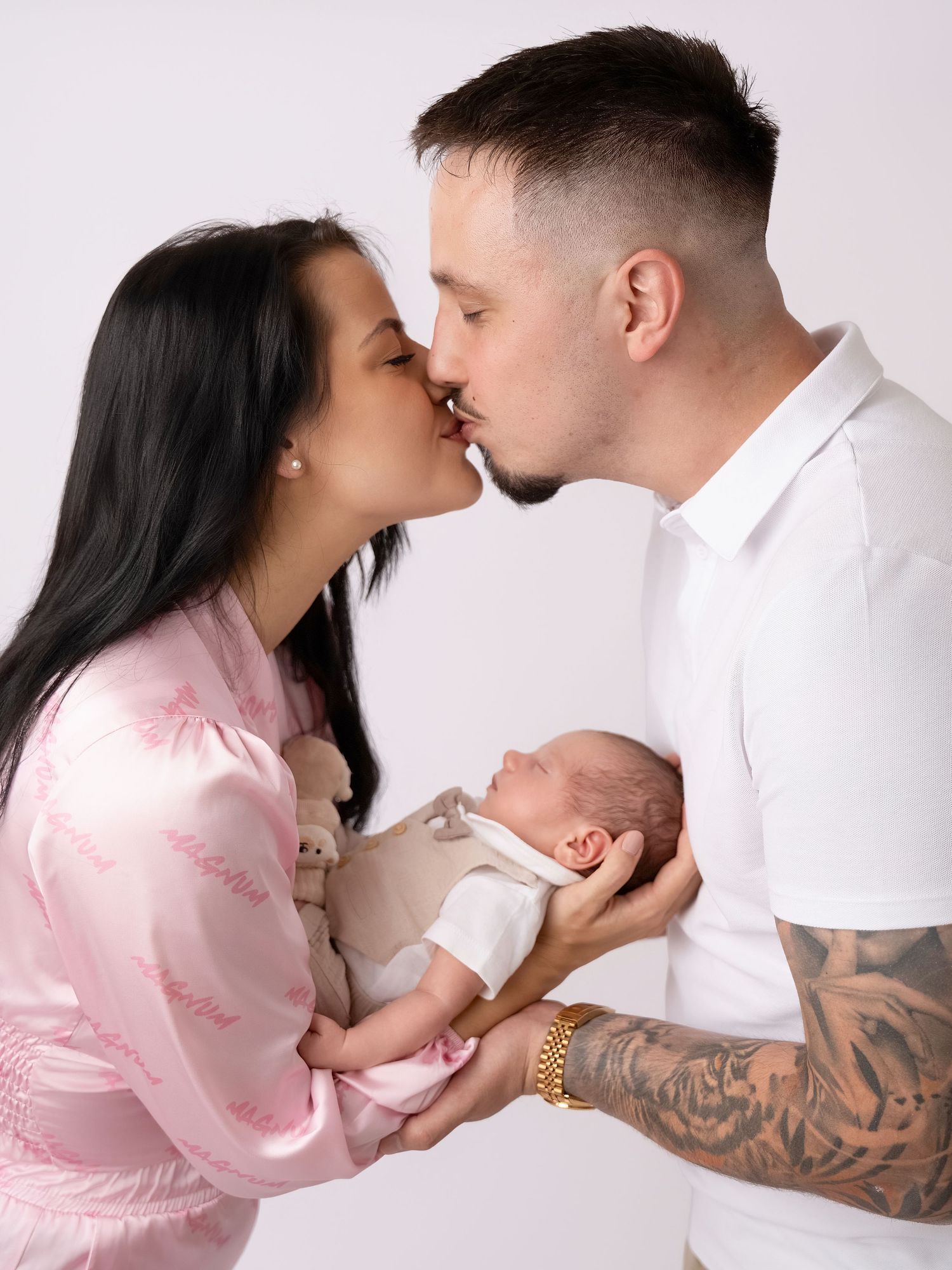 A couple shares a kiss while holding their newborn baby, the woman in pink satin and the man with tattoos, on white background.