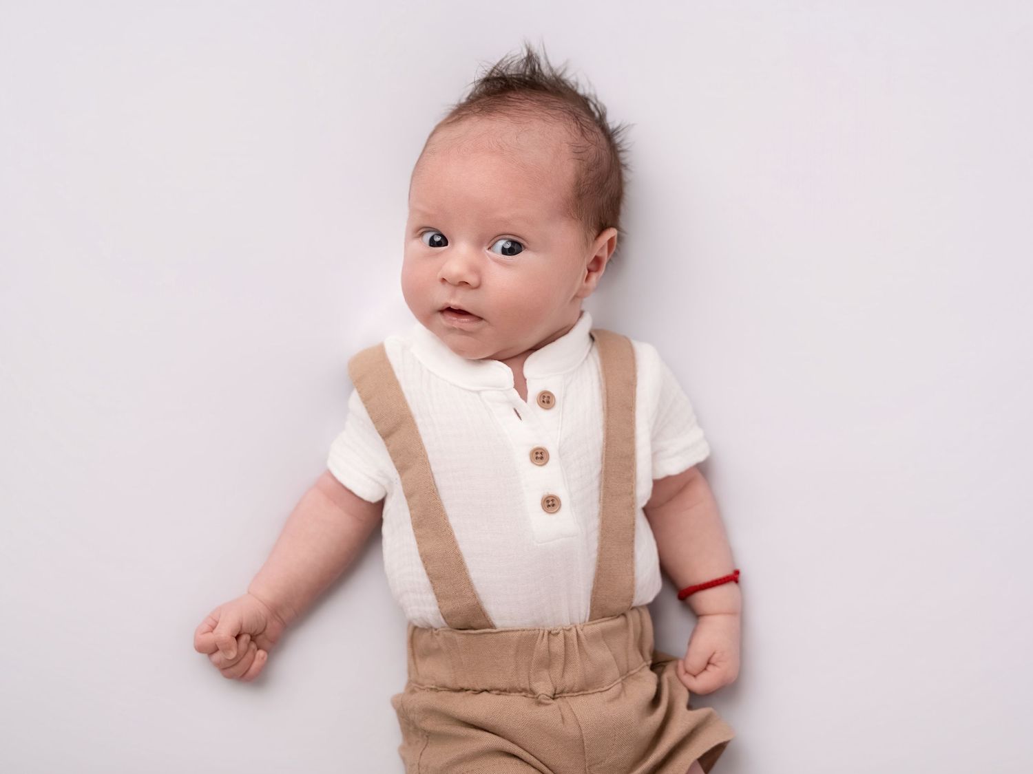 Adorable baby boy in white polo shirt and tan suspender shorts lying on a light gray background.