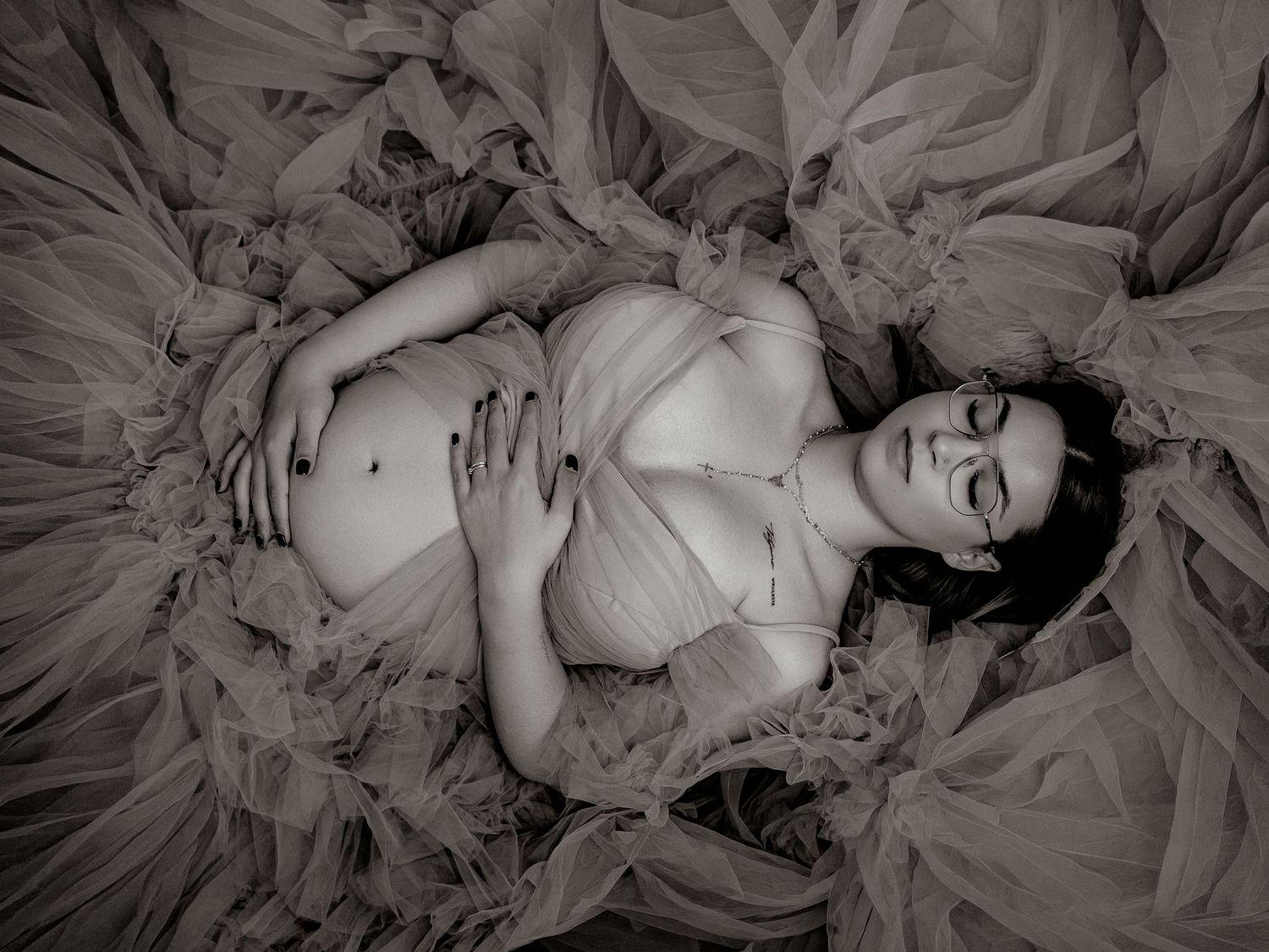 Black and white maternity photo of a pregnant woman lying in flowing tulle fabric, cradling her belly.