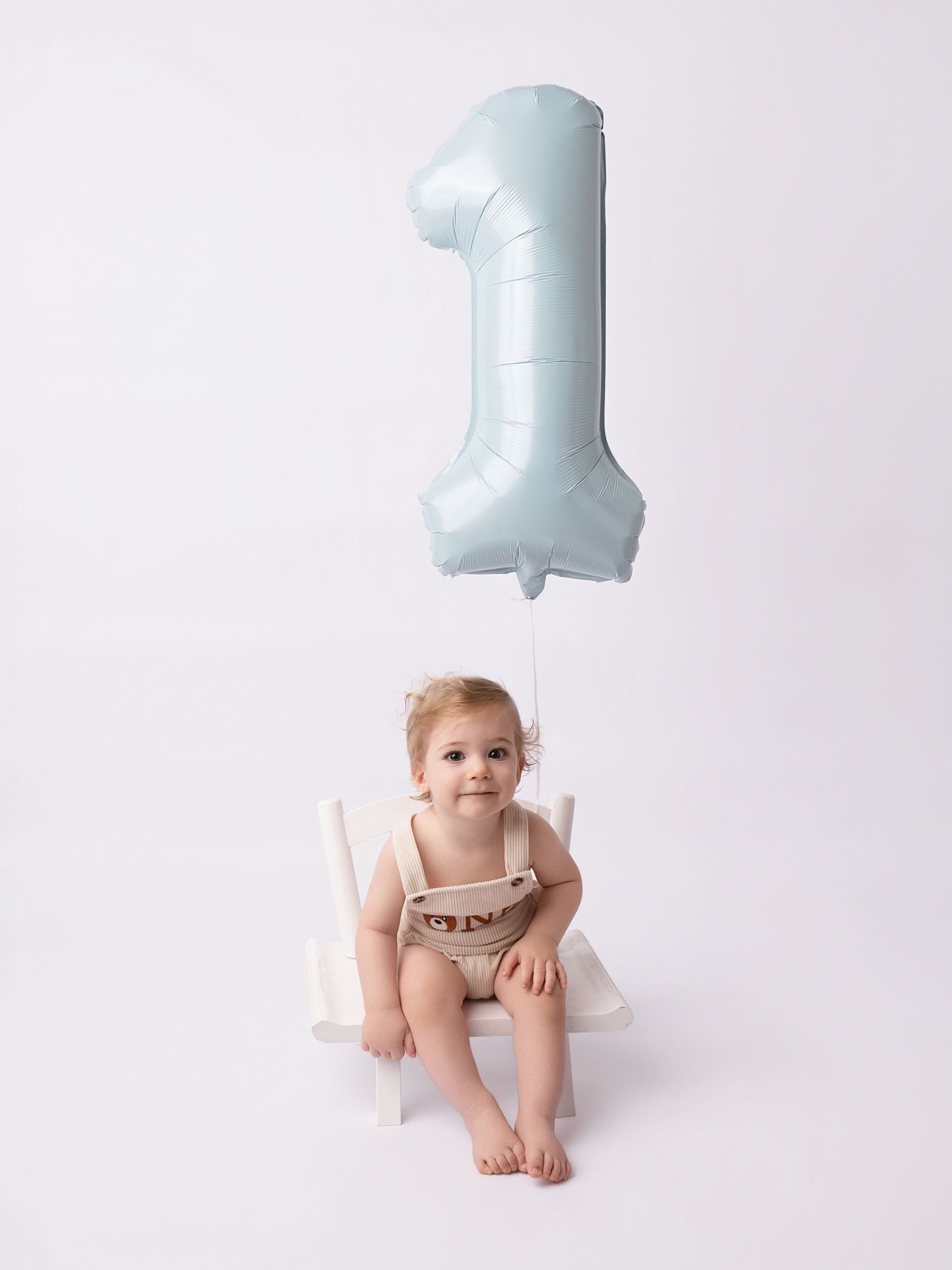 Baby sitting on white chair with light blue number one balloon for first birthday photo shoot.