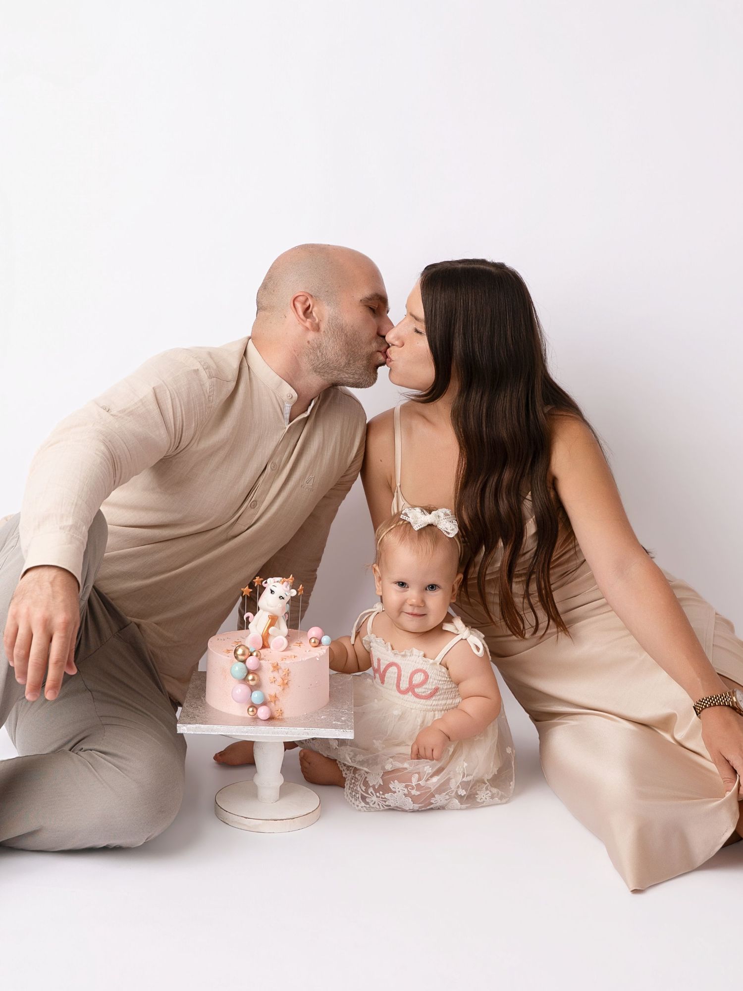 Parents kiss behind baby girl celebrating first birthday with pink giraffe smash cake in studio.