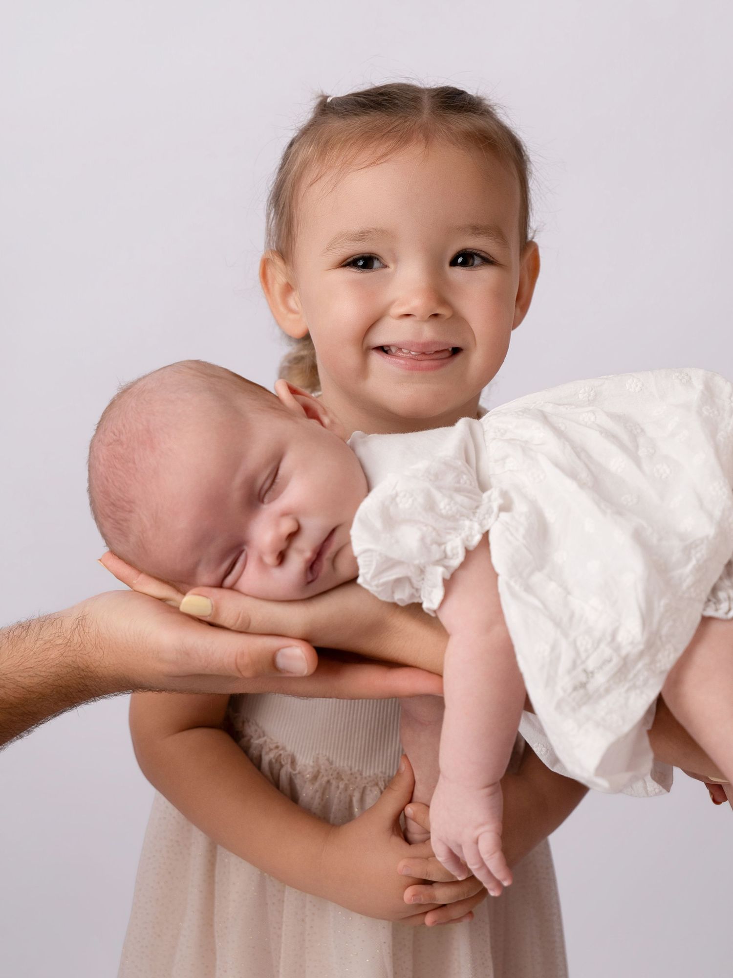 Smiling toddler girl holds sleeping newborn baby sibling, supported by adult hands, in studio photo.