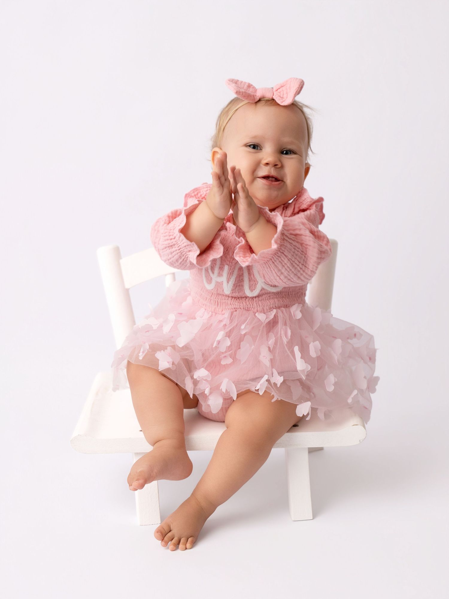 Baby girl in pink ruffle dress and bow headband clapping hands, seated on white chair for milestone photo.