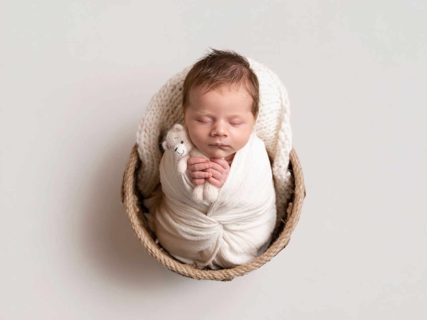 Peaceful newborn baby swaddled in cream wrap sleeping in a round wicker basket with a small white toy.