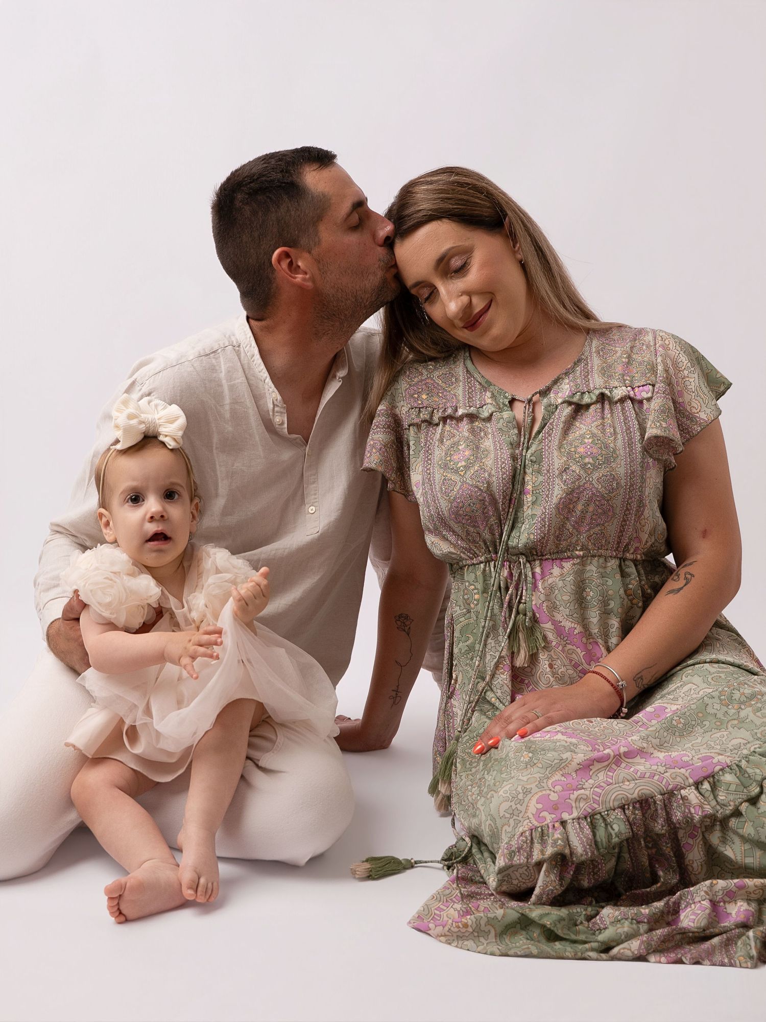 A family portrait with a man kissing a woman's cheek as they sit with their baby girl in a white studio.