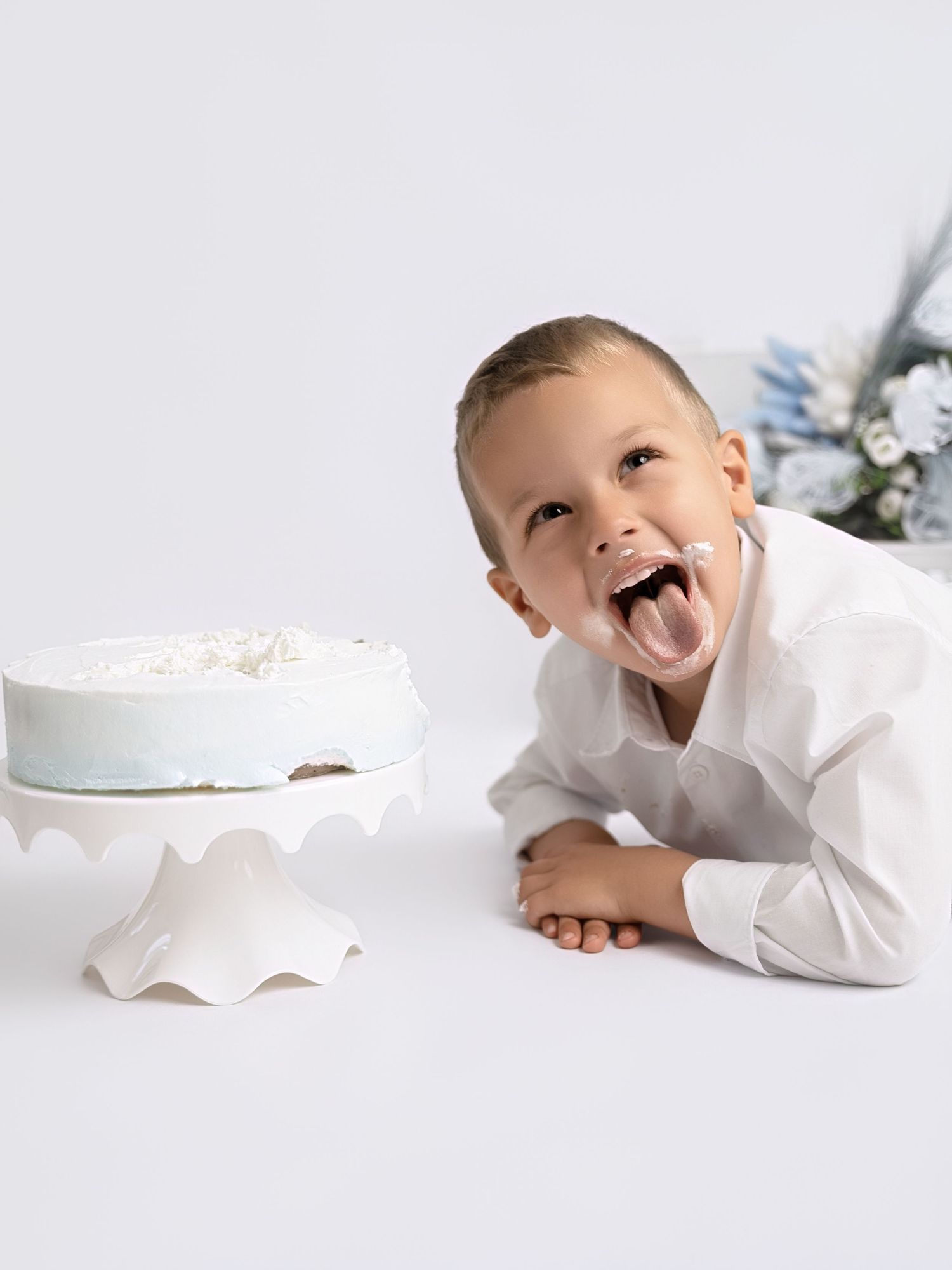 Young boy in white shirt laughs with tongue out beside white frosted birthday cake on pedestal.