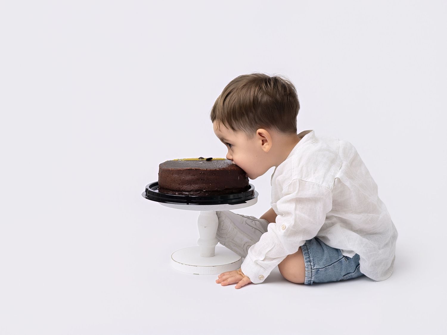 Toddler boy in white shirt sitting on floor reaching toward chocolate cake on white pedestal stand.