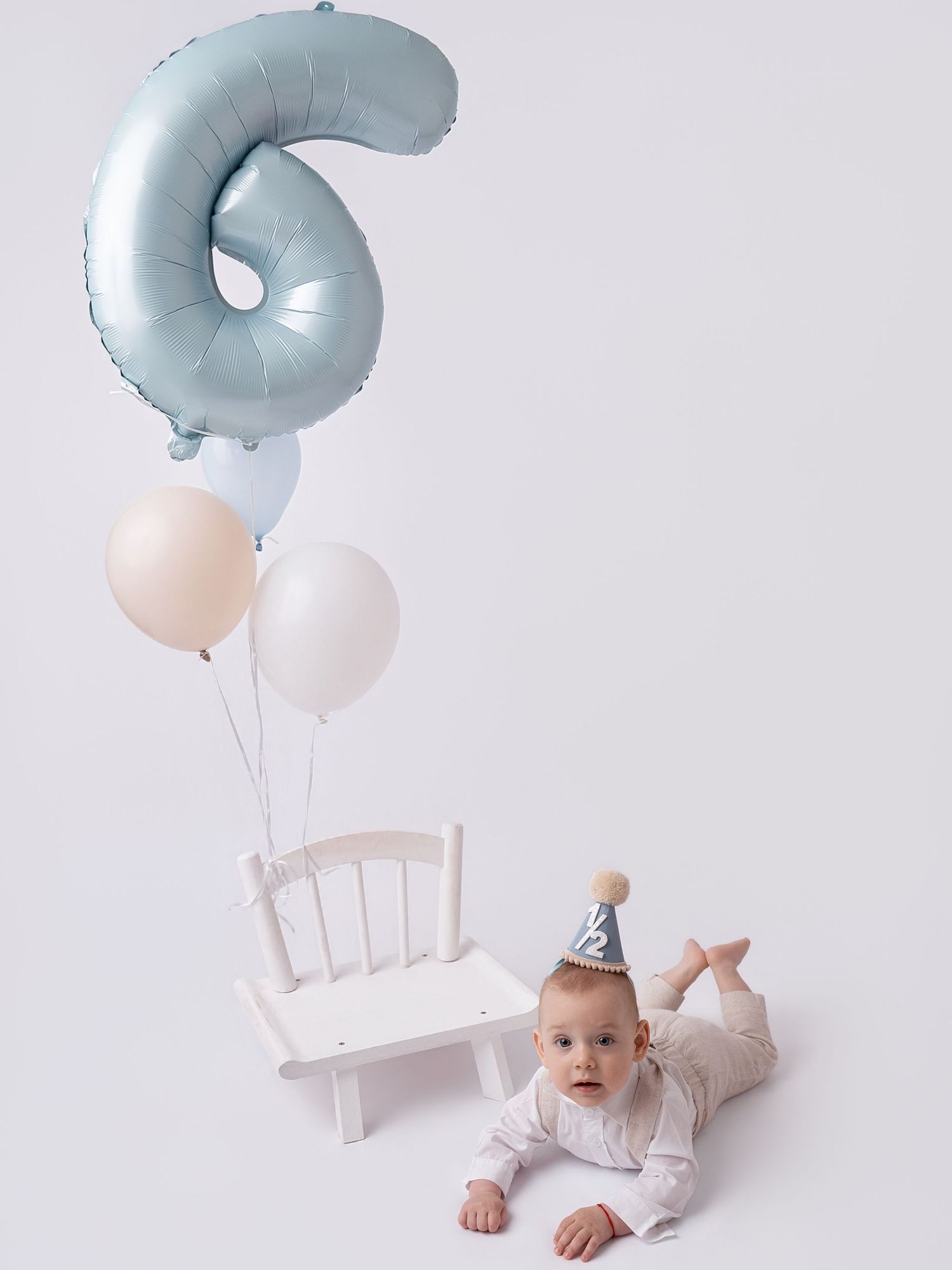 Adorable baby wearing silver party hat poses with blue number 6 foil balloon and white chair for birthday photo.