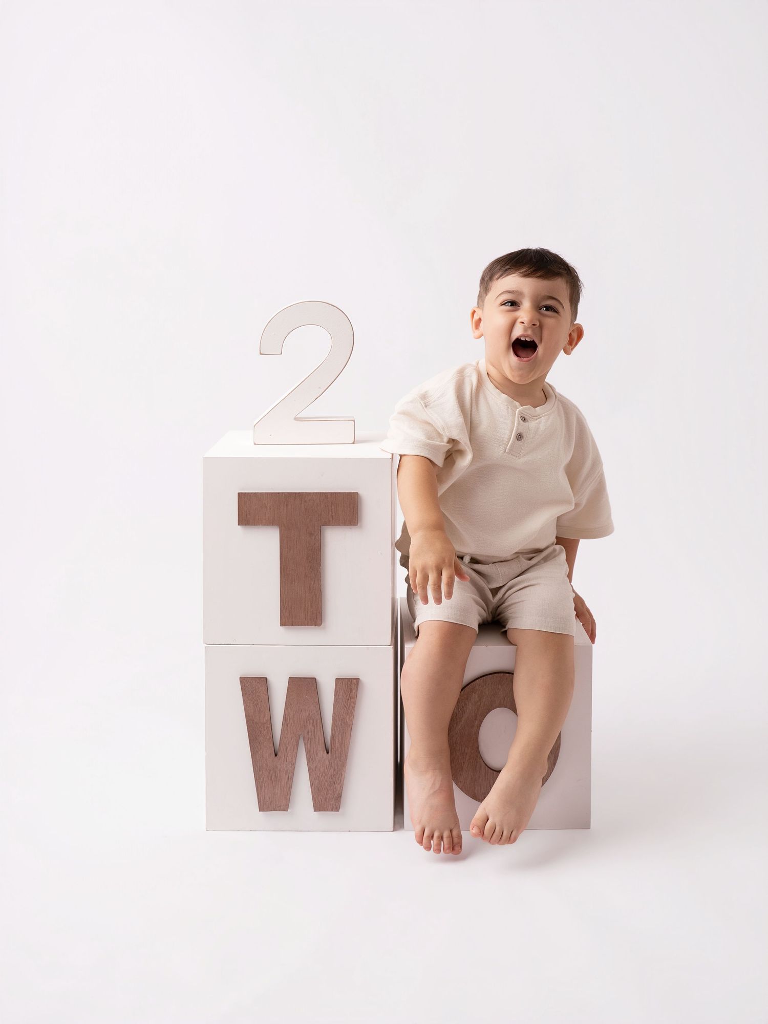 Laughing toddler boy sitting on white alphabet blocks spelling TWO for a 2nd birthday photo session.
