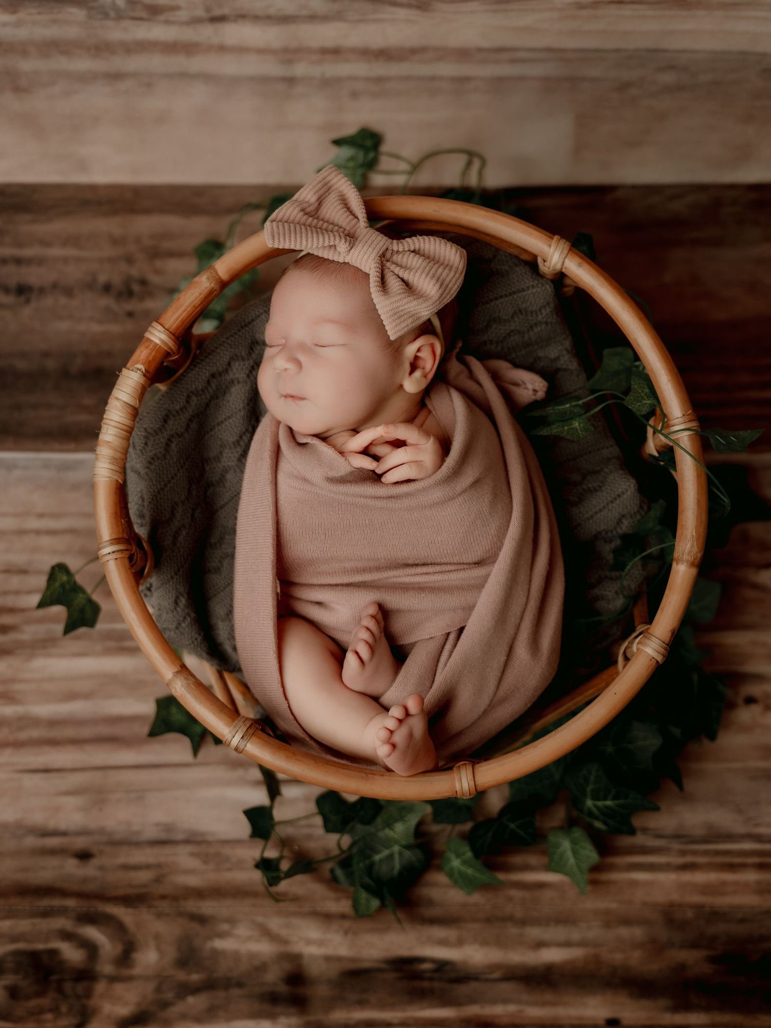 Newborn baby girl swaddled in pink wrap with bow headband sleeping peacefully in a wicker basket with ivy.