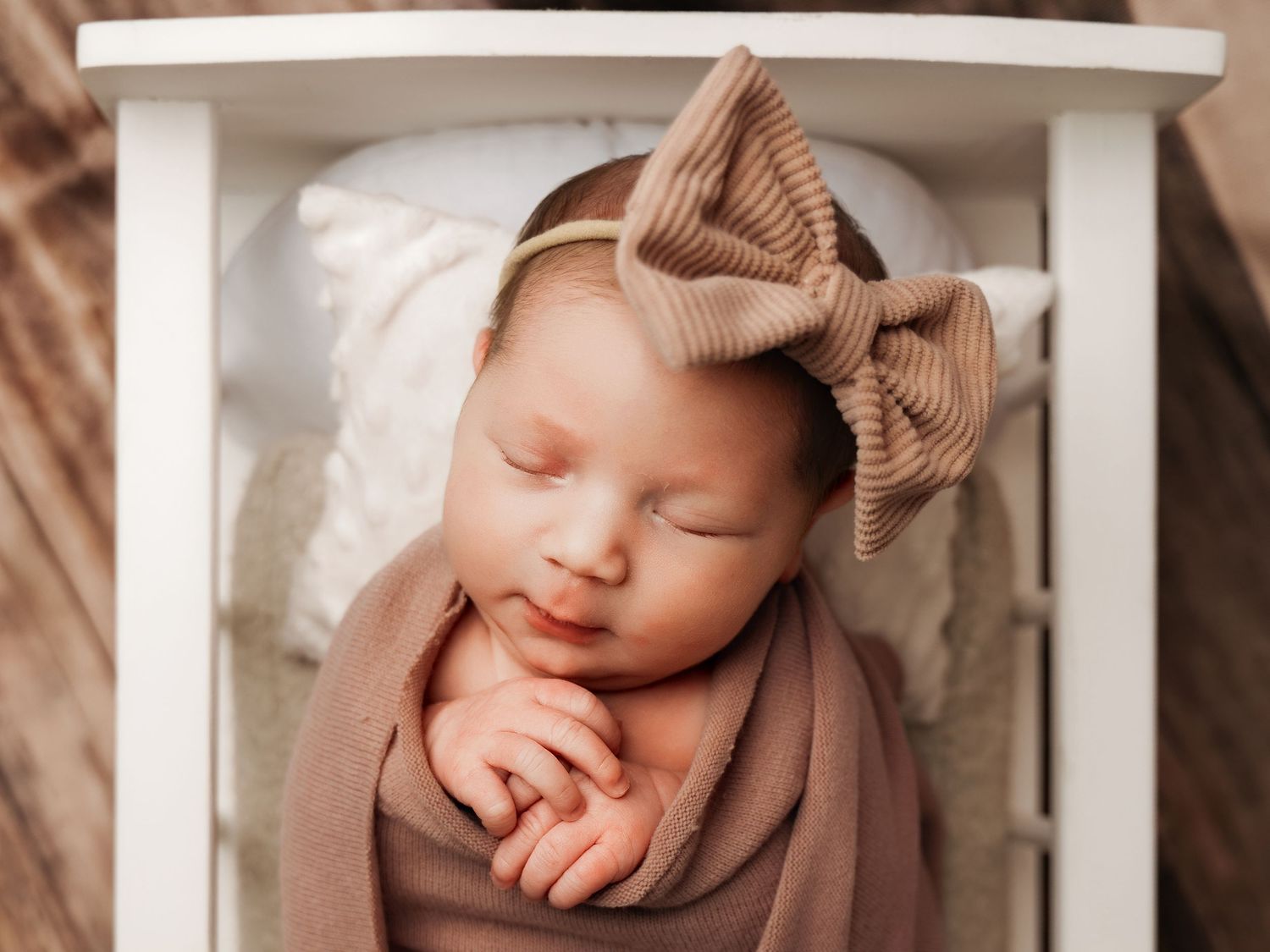 Sleeping newborn baby wrapped in mauve swaddle with tan bow headband in white mini crib on wood backdrop.